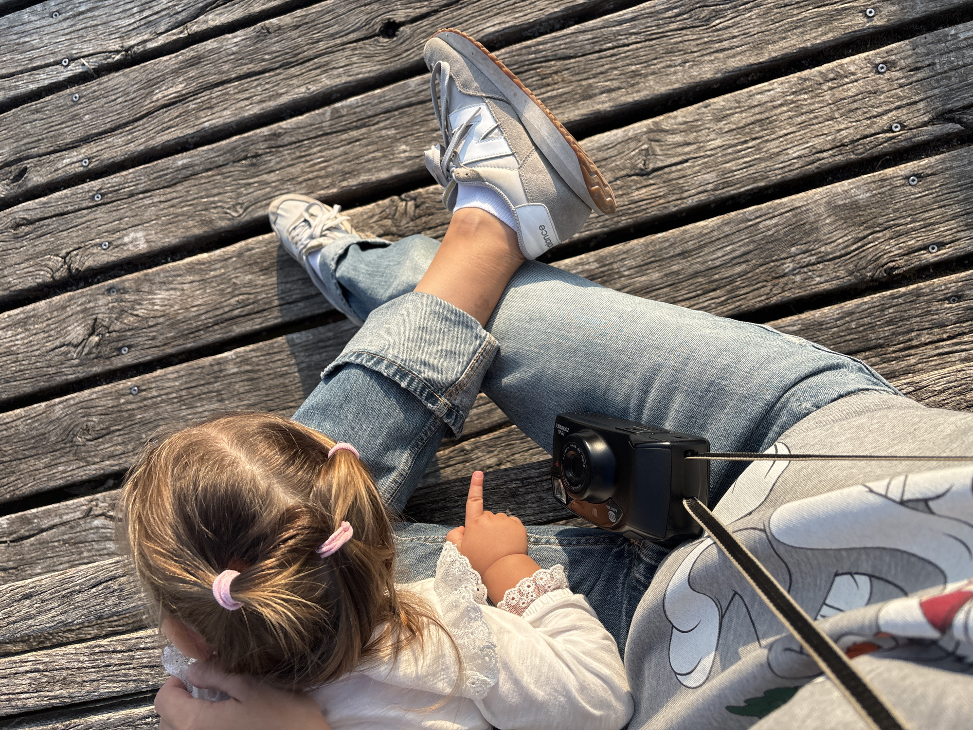 An overhead view shows a person sitting on a wooden pier, possibly in Bad Buchau, Germany, with a young child. The child, wearing a white lace-trimmed shirt and blue jeans, has their hair in two pigtails secured with pink ties. The child's left hand is extended, pointing towards a black digital camera positioned on the pier next to them. The person's legs, also in blue jeans, are bent, and they are wearing grey and white athletic shoes. The wooden planks of the pier are weathered and show visible grain and nails. The lighting suggests it is late afternoon.