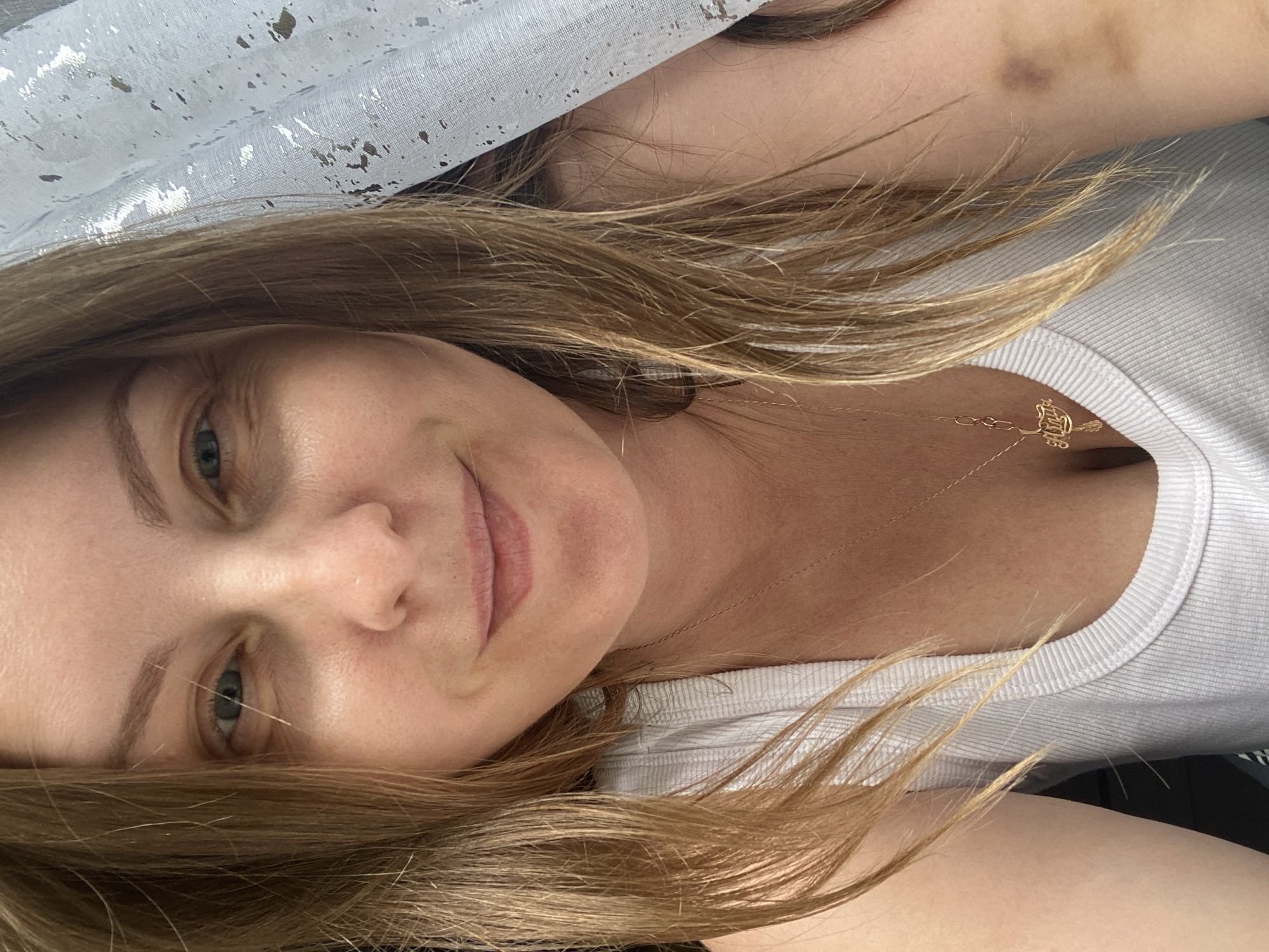 The image shows a close-up of a woman's face and upper chest, taken from a high angle. The woman has light brown hair, fair skin, and blue eyes. She is wearing a white tank top and a gold necklace with a pendant. Her hair is partially covering her face and is falling to her left. To the right of the woman, a white curtain with silver speckles is visible, obscuring part of the background. A bruise is visible on the woman's upper arm, near her shoulder. The location in the city of Noviy, Russia is not specified or discernible from the image.