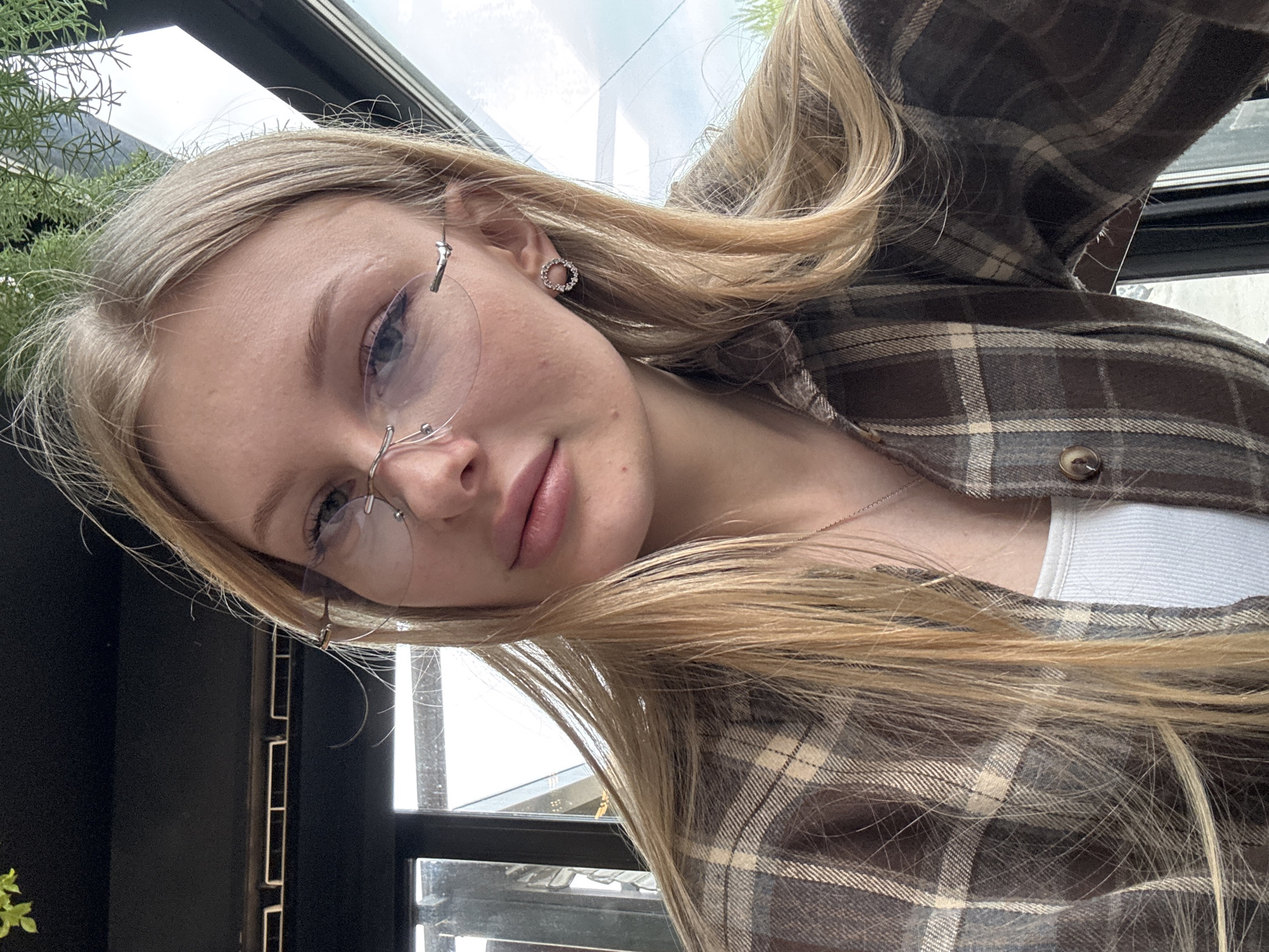 A young blonde woman is pictured from a low angle, wearing rimless eyeglasses and small circular earrings. She is dressed in a brown and tan plaid flannel shirt over a white undershirt. The setting appears to be indoors near a window. Some greenery is visible near the top left corner of the image. The location of the image is not revealed.