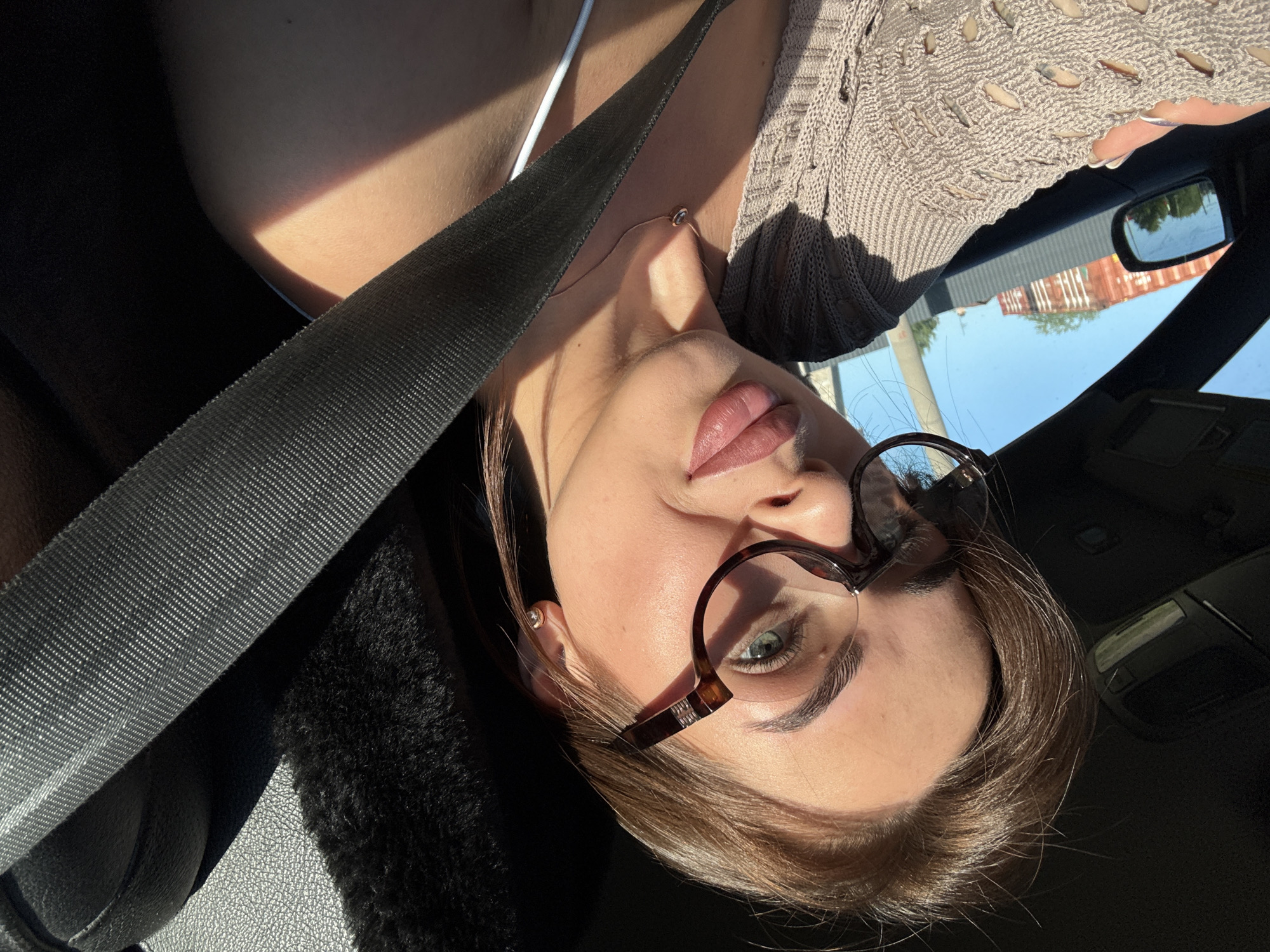 A young woman is photographed from a low angle inside a car. She is wearing a seatbelt, eyeglasses, and a beige knit sweater. She has fair skin, full lips, and light brown hair. The background shows the car interior, a side mirror reflecting buildings. The location is likely Perm, Russia, due to the buildings' style and the presence of what appears to be Russian industrial architecture in the reflection.