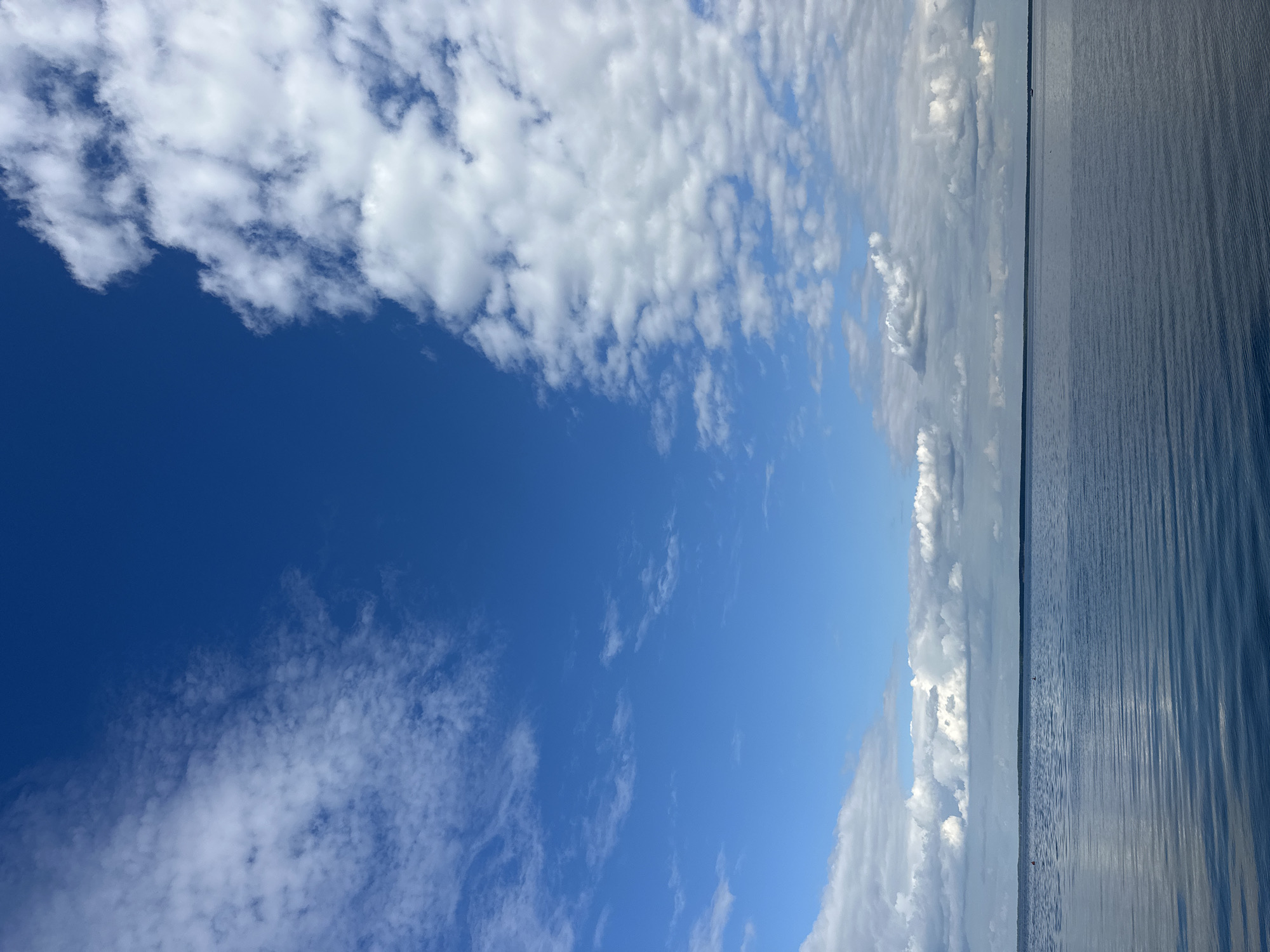 A wide, still body of water stretches across the lower half of the frame, reflecting the blue sky and scattered white clouds. The water's surface is mostly calm, with subtle ripple patterns creating gentle lines. A dark, narrow strip of land, likely a treeline, forms the distant horizon. Above, a vast expanse of bright blue sky dominates, punctuated by fluffy white clouds that range from thin wisps to more substantial formations. The scene evokes a sense of openness and tranquility. This appears to be a natural landscape, potentially a lake or a large river, located in the Нарацкі сельскі Савет, Belarus. No people or specific man-made structures are clearly visible in this image, suggesting a natural or remote setting.