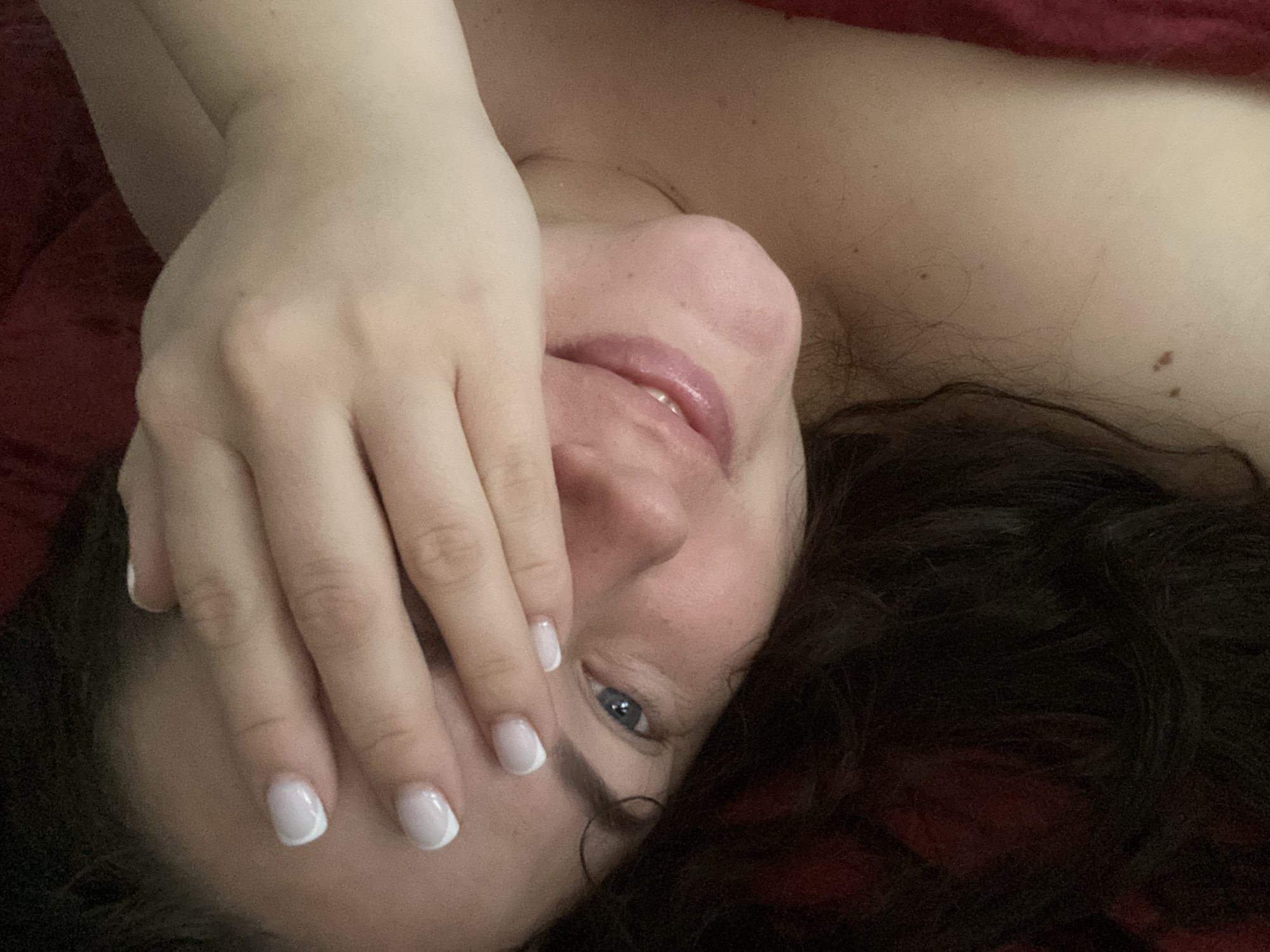 A woman with dark hair and fair skin is lying on a red surface, possibly a bed or blanket. Her face is partially covered by her hand. The visible eye is blue. Her nails are manicured with a white crescent design. It is impossible to determine the location is Sochi, Russia based solely on this image.