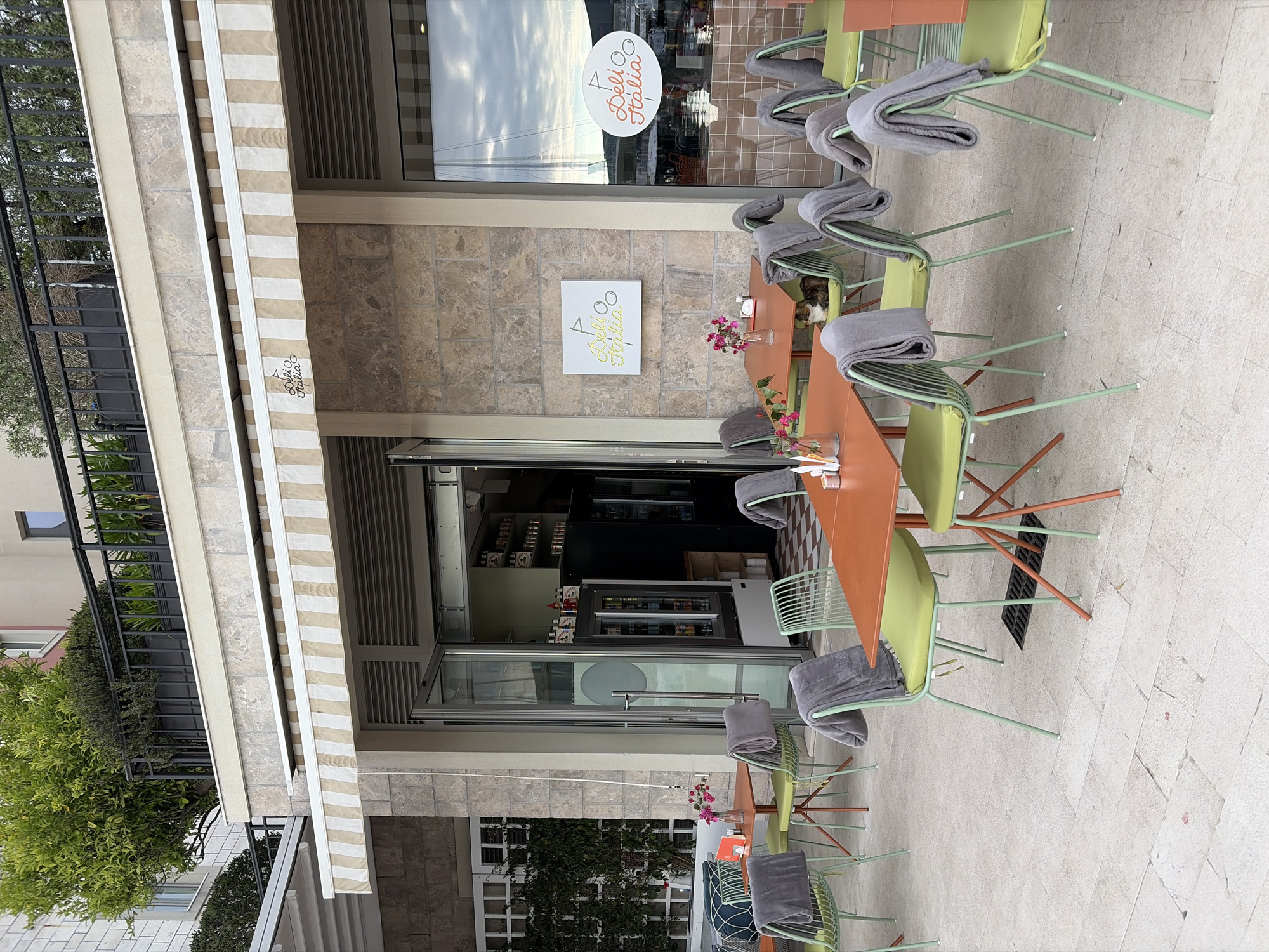 The image shows the exterior of a café called "Deli Italia" in Tivat, Montenegro. The café has an open doorway leading to an interior with shelves and refrigerated display cases. Outside, there are several tables and chairs arranged. Each chair has a folded gray blanket on it. Small vases with flowers are placed on some of the tables. An awning with beige and white stripes extends over the front of the café. The building facade is made of stone. A logo for "Deli Italia" is visible on the window and on a sign on the wall. Vegetation is present to the left and right of the building.