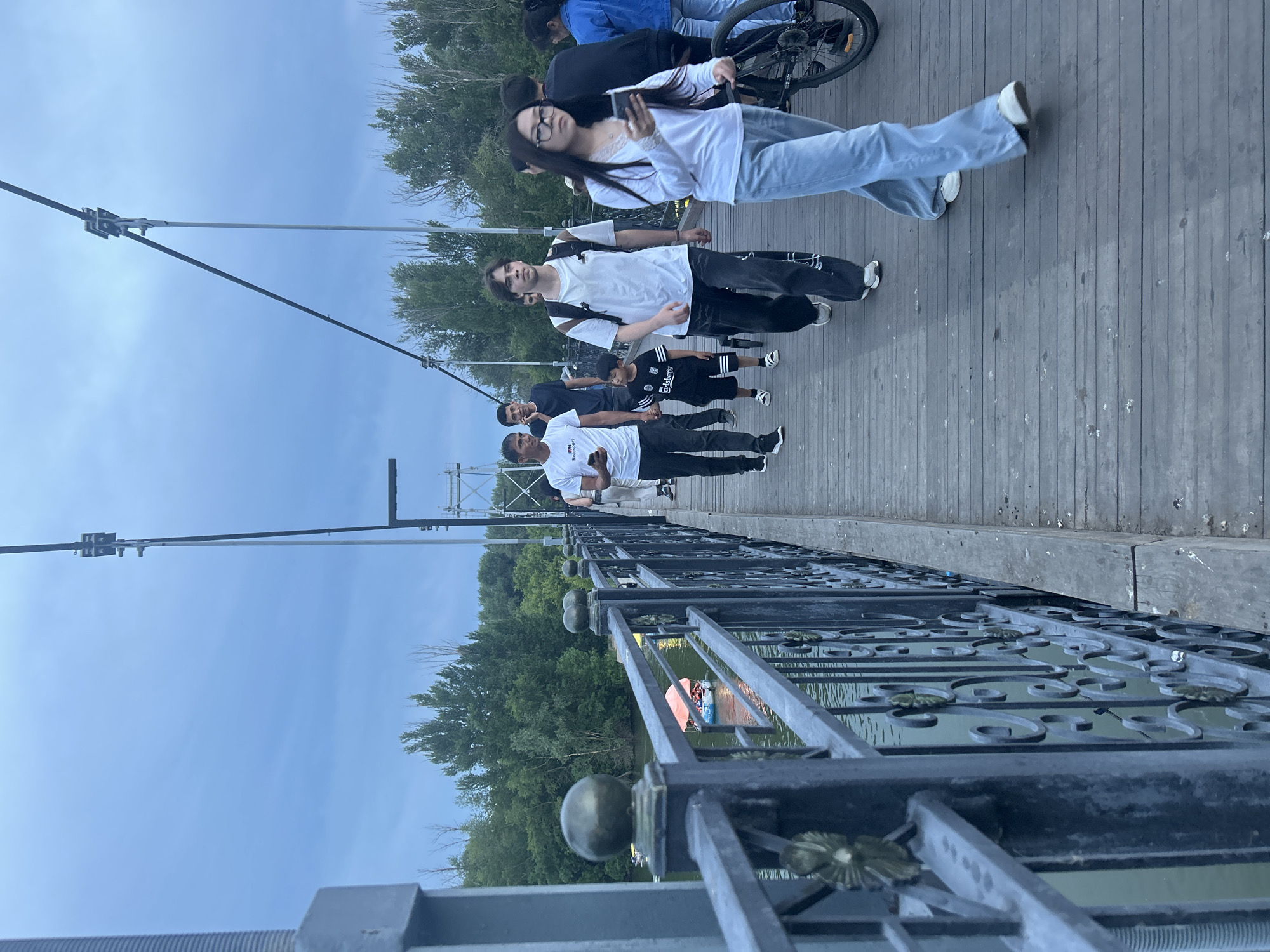 On an overcast day, pedestrians stroll across a wooden suspension bridge in the city of Oral, Kazakhstan, with a calm body of water visible below and lush green trees lining the banks. The bridge features a decorative metal railing. Among the individuals are a young boy in a black and white striped t-shirt and shorts, a young man with a backpack, a woman with glasses looking at her phone, and others walking in both directions. A bicycle is also present on the bridge. In the water below, a small boat with an orange canopy is being pedaled by passengers. The overall atmosphere appears relaxed, with people enjoying a walk over the river.