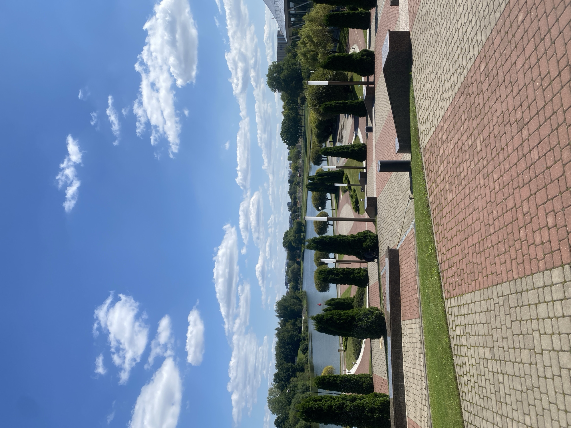 The image depicts a scenic view of the Svislach River embankment in Minsk, Belarus, on a clear, sunny day. The foreground is dominated by a paved walkway with a pattern of red and grey rectangular stones, bordered by a strip of green grass. Several tall, conical evergreen trees are strategically placed along the walkway, interspersed with modern lampposts. In the midground, the calm Svislach River stretches out, reflecting the blue sky dotted with fluffy white clouds. The riverbank is lined with lush green trees and some bushes, creating a verdant backdrop. Across the river, more trees and buildings are visible, suggesting a park or a more urbanized area in the distance. On the right side of the image, a modern building with a large, slanted roof and glass facade is partially visible, indicating a contemporary architectural presence. There are no visible people in the image, contributing to a sense of tranquility and stillness. The overall impression is one of a well-maintained public space designed for relaxation and enjoyment of nature within an urban setting.