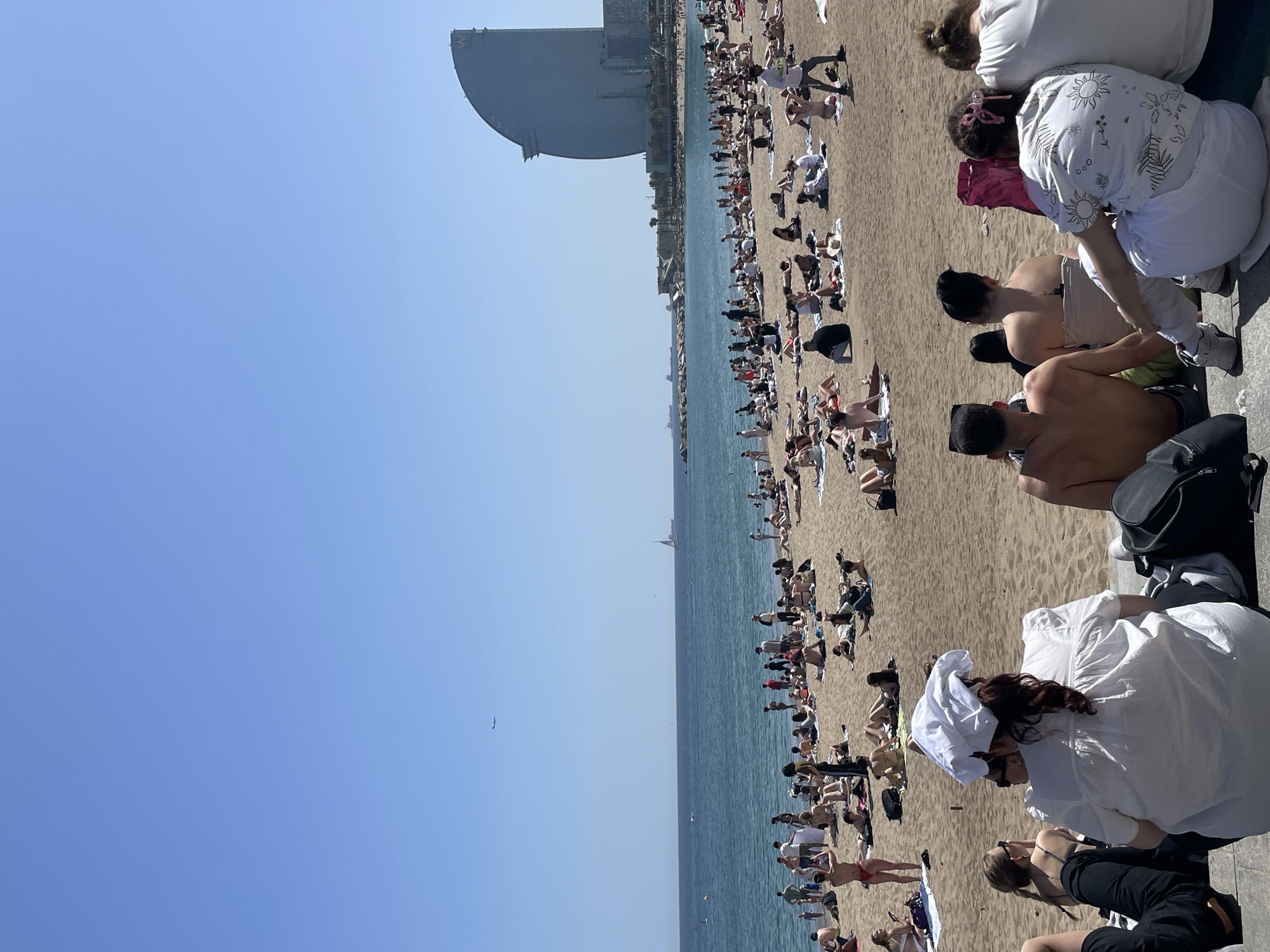 The image shows a crowded beach on a sunny day. The beach is located in Barcelona, Spain, and the Hotel W Barcelona can be seen in the distance. People are sunbathing, swimming, and walking along the shore. Many of the people are scantily clad.
