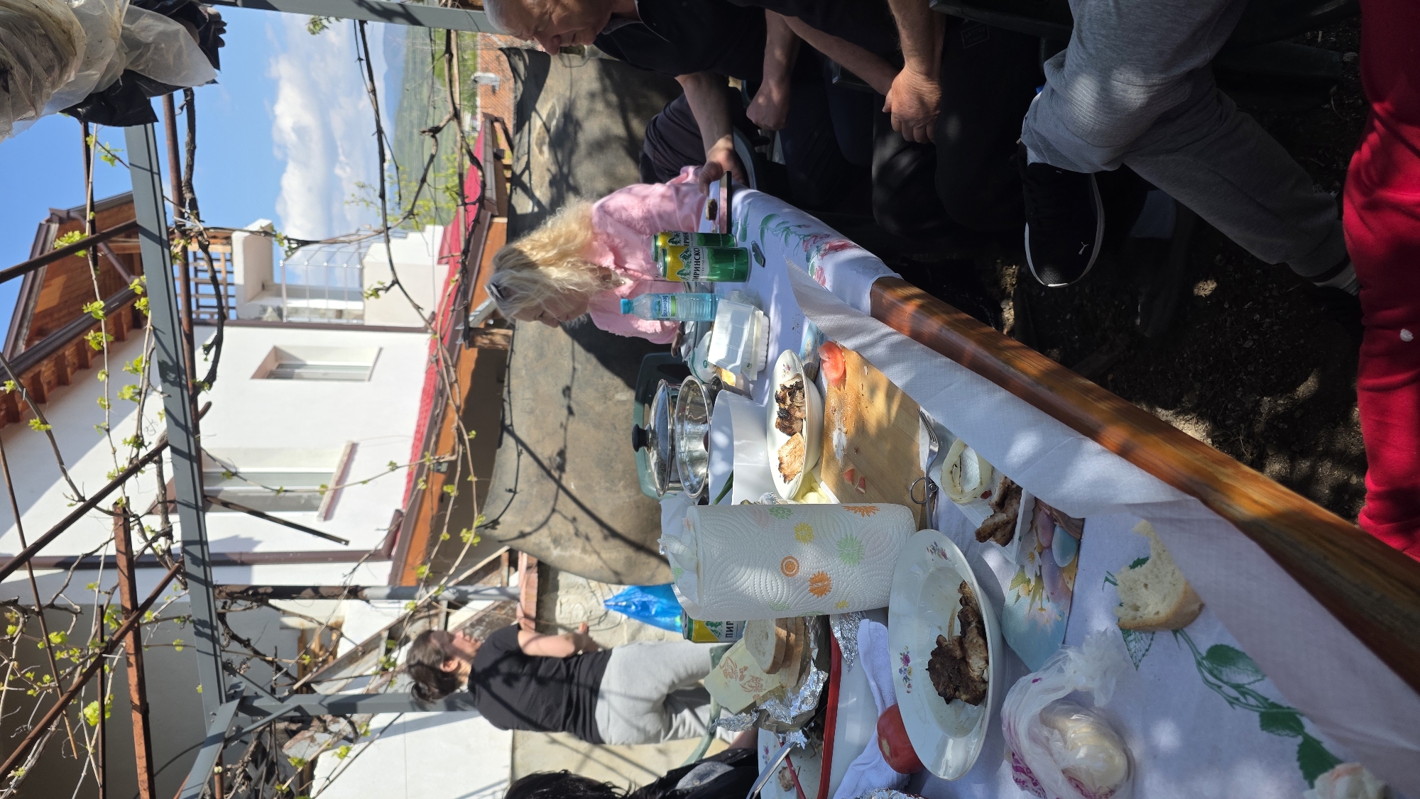The scene depicts an outdoor gathering in what appears to be a backyard setting in Katuntsi, Bulgaria. A long table covered with a white tablecloth holds an assortment of food items, including plates of meat, bread, tomatoes, and paper towels. People are gathered around the table, including one woman wearing a pink shirt who appears to be interacting with the food and beverages. Several other people are seated or standing near the table. The background includes a white house with visible windows and a reddish-brown roof. A vine structure partially obscures the view, and the sky is visible with some clouds. The presence of food, beverages, and people suggests a meal or celebration taking place outdoors.