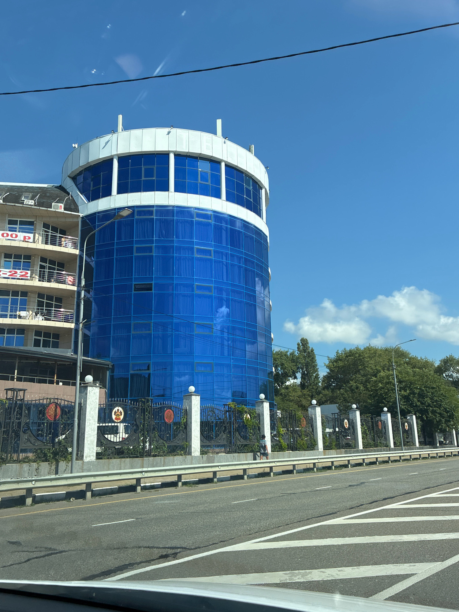 This image shows a modern, cylindrical building with a bright blue glass facade, situated in the city of Lermontovo, Russia. The building is multi-storied and features white accents around its top and windows. To the left of the blue building, a lighter colored, multi-story building with balconies and signage is visible. A dark metal fence with decorative elements and what appear to be crests or emblems separates the buildings from a road. The road in the foreground has lane markings and a metal guardrail. Trees and lush greenery are visible behind the fence, and the sky is a clear, vibrant blue with a few scattered white clouds. A power line stretches diagonally across the top of the frame. A person is seen walking on the sidewalk behind the fence. The overall scene suggests an urban environment with modern architecture, possibly a commercial or administrative area.