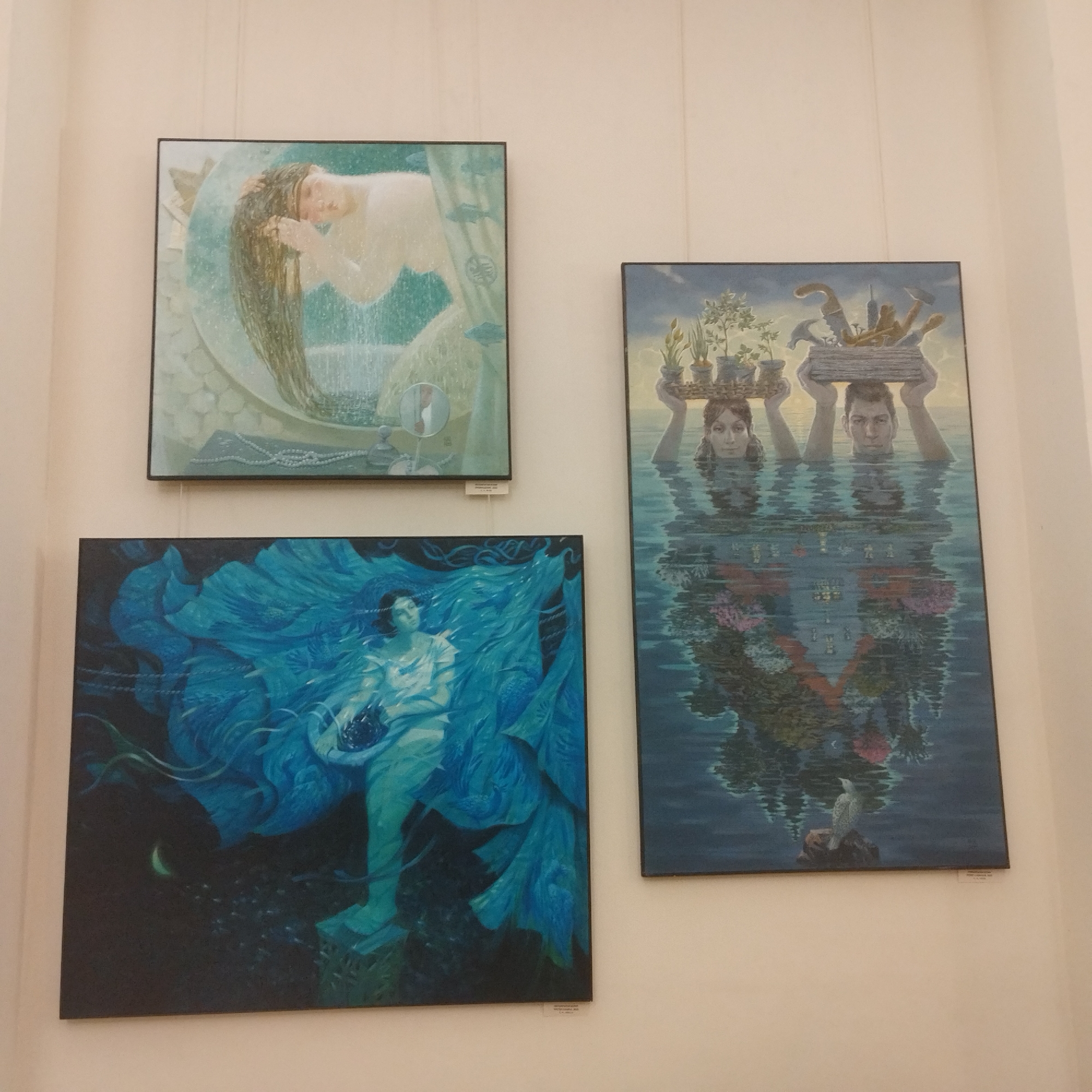 The image shows a wall in a gallery or exhibition space in Yekaterinburg, Russia, displaying three framed paintings.
The painting on the upper left depicts a woman with long hair lying down in a crescent-shaped container. The background is a light green color with what looks like running water.
The painting on the lower left features a woman surrounded by large blue petals or flower-like forms in a dark setting, possibly at night, as indicated by a crescent moon in the corner.
The painting on the right shows a man and a woman immersed in water up to their necks. They are both holding up a flat object containing plants and tools. The water reflects a building or structure with flower decorations, and a bird is perched on a rock in the lower part of the image.