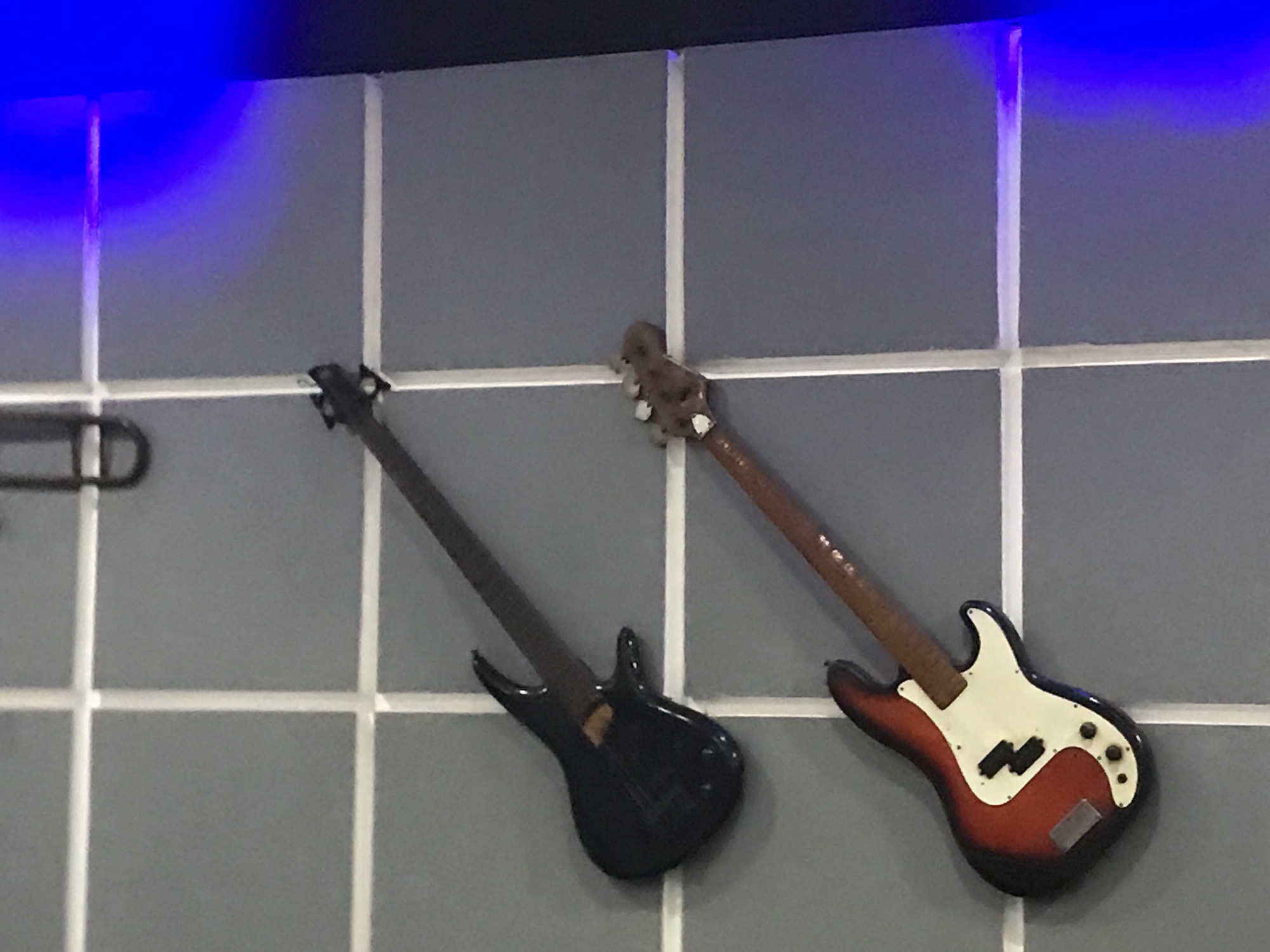 The image shows two miniature electric bass guitars mounted on a tiled wall. The tile grout lines are white, while the tiles themselves are gray. The guitar on the left is black, and the guitar on the right is red, white, and black. A small portion of a black coat hanger is visible on the left side. A blue light source is visible in the upper left corner of the image. The photo was taken indoors in the City of Medan, Indonesia.