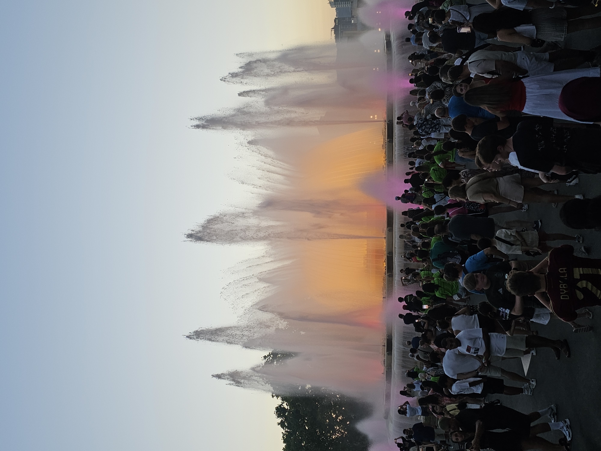 The image captures a crowd of people gathered in front of a large, illuminated fountain in Barcelona, Spain. The Magic Fountain of Montjuïc is the central subject, with water jets reaching upwards and illuminated by pink and orange lights. The sky above is a gradient of light blue, suggesting it is either dawn or dusk. Many individuals in the crowd are holding up phones, seemingly to take pictures or record the spectacle. The people are dressed in casual summer attire. Some are standing close to the fountain, while others are further back, creating a dense gathering. The fountain is set in a tiered structure, with the water flowing down into lower levels. In the background, trees and some distant buildings are visible.