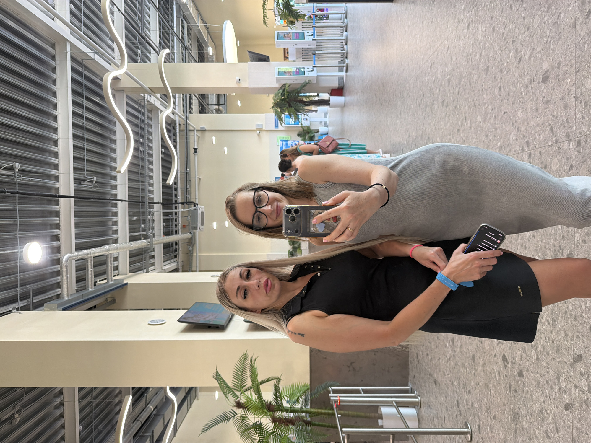 Two women are posing for a photo in what appears to be the International Airport Sheremetyevo in Moscow, Russia. The woman on the top is wearing glasses, a light-colored dress, and holding a phone. The woman on the bottom is wearing a black dress and holding a phone as well. Behind them, there are palm trees and what appear to be check-in kiosks. The ceiling has an industrial design with visible ductwork and wavy lighting fixtures.