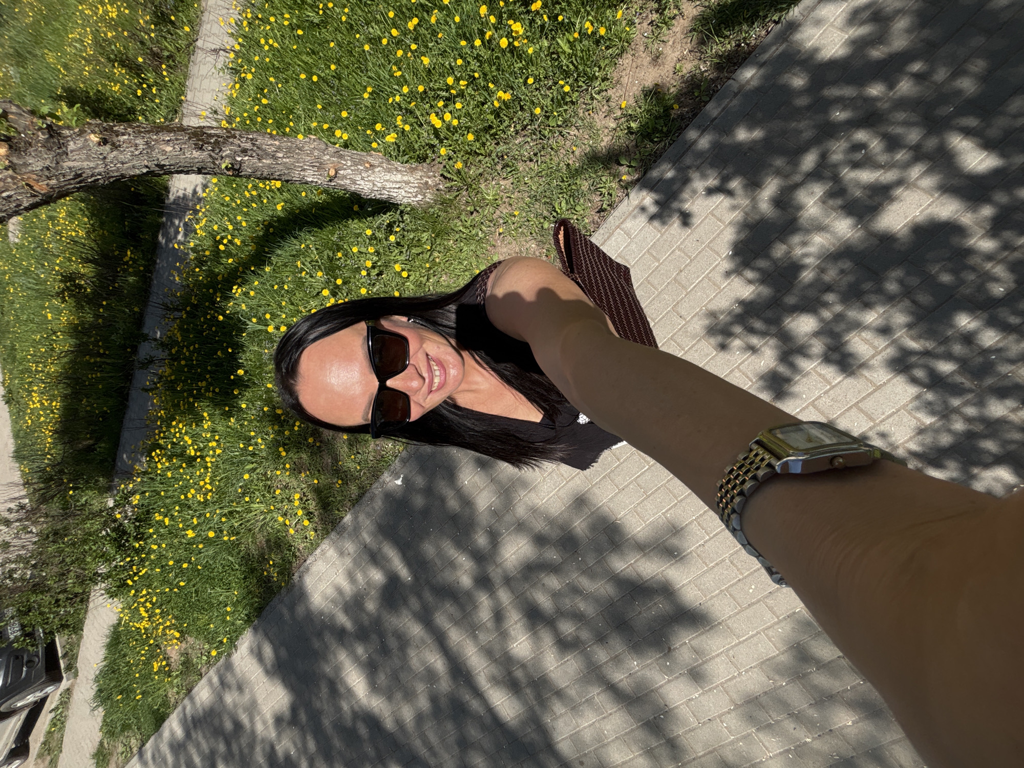 A woman with long dark hair and sunglasses is taking a selfie outdoors on a sunny day. She is wearing a dark top and holding a brown woven bag on her left shoulder. On her right wrist, she is wearing a metallic watch. She is standing on a paved sidewalk, and the shadows of trees fall across the pavement. To the left of the sidewalk, there is a grassy area covered with yellow dandelions and a tree branch extending over the grass. In the distance, a vehicle is partially visible. The location is likely the city of Chaikovsky, Russia.