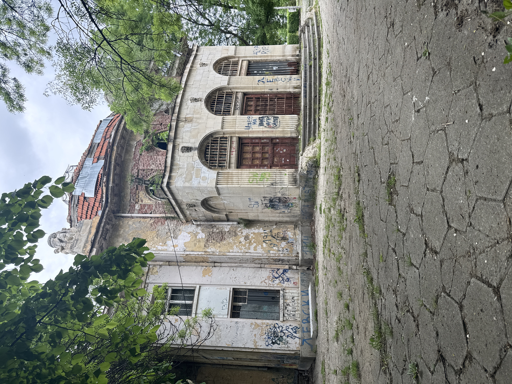 The image shows an abandoned, derelict building in Sofia, Bulgaria. The structure has a neoclassical architectural style with arched windows and decorative columns on the upper level. The lower portion is constructed of brick and decaying white plaster. The building is covered in graffiti. The roof is partially tiled and partially covered with metal sheeting. A stone walkway leads up to the entrance, and vegetation grows around the base of the building and along the path. The sky is overcast, and trees surround the building. There are no people visible in the image.