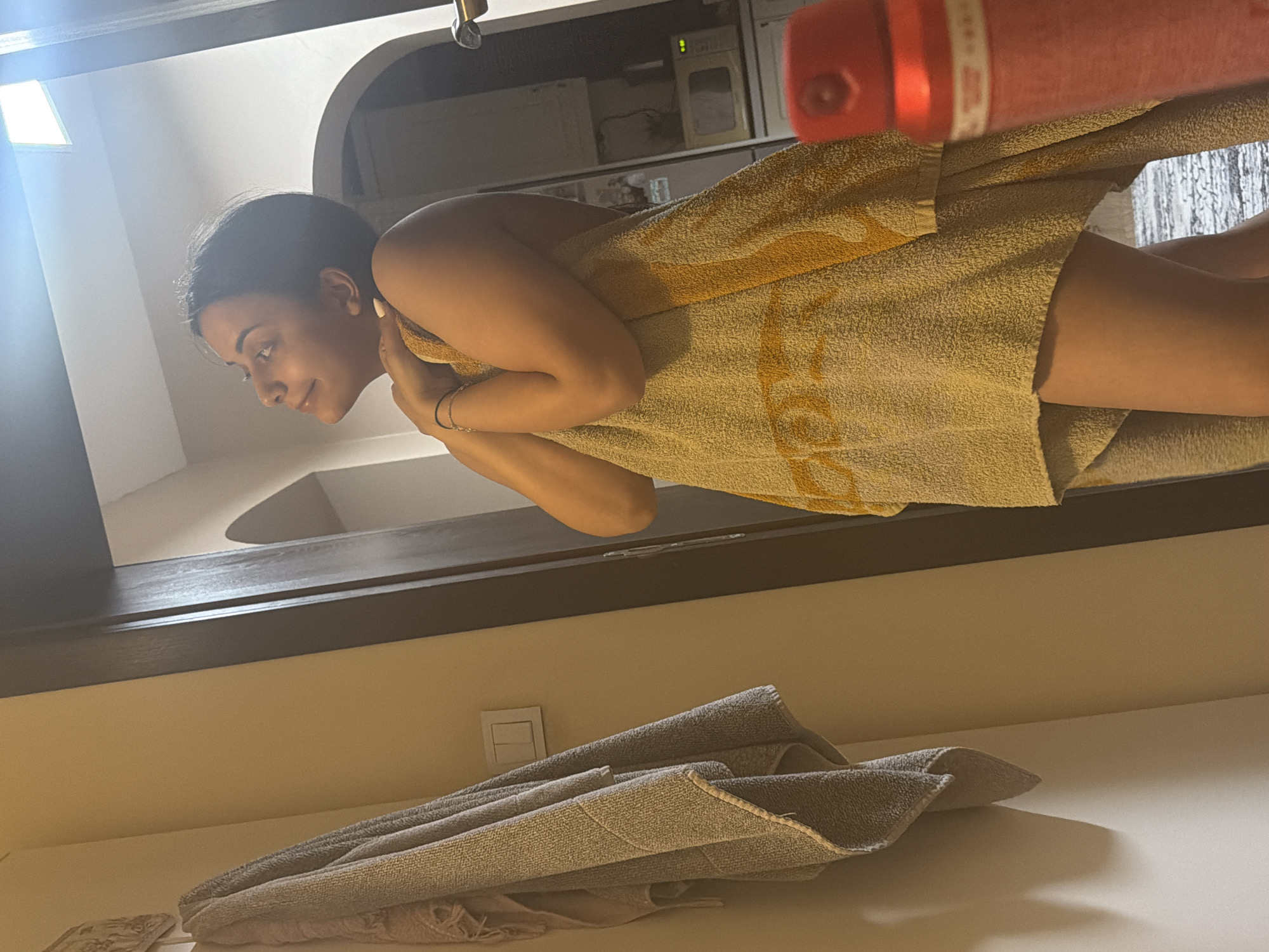 The image captures a young woman, likely in her 20s or 30s, wrapped in a large, light-colored towel, possibly beige or yellow, with a subtle pattern. She is seen in profile, looking towards the left with a gentle smile, suggesting a relaxed or contented mood. Her dark hair appears to be pulled back. She wears a thin bracelet on her left wrist, and her fingernails are painted a light, possibly white or cream, color.
The photograph is taken from a low angle and is rotated ninety degrees counter-clockwise. If viewed upright, the woman would be standing, with a dark wooden or metal beam/frame forming a horizontal barrier below her. Above this beam, behind the woman, the background features white walls, a bright light source on the left, and an arched opening on the right. Further back, some indistinct appliances or shelves with a green indicator light are visible. A red object, possibly a spray bottle or container with some blurred white text (perhaps "WHITE" or "WATER") on it, is partially visible in the upper right corner of the original orientation (upper left when upright).
In the foreground of the original image (below the dark beam), several folded grey towels are stacked on a flat surface, which appears to be a counter or table. A white light switch is also visible on the wall in this lower section.
The overall setting appears to be indoors, likely a bathroom or a spa, given the presence of towels and the woman's attire. There are no specific visual cues within the image to indicate its location as Shumen, Bulgaria. The time of day cannot be determined from the indoor lighting.