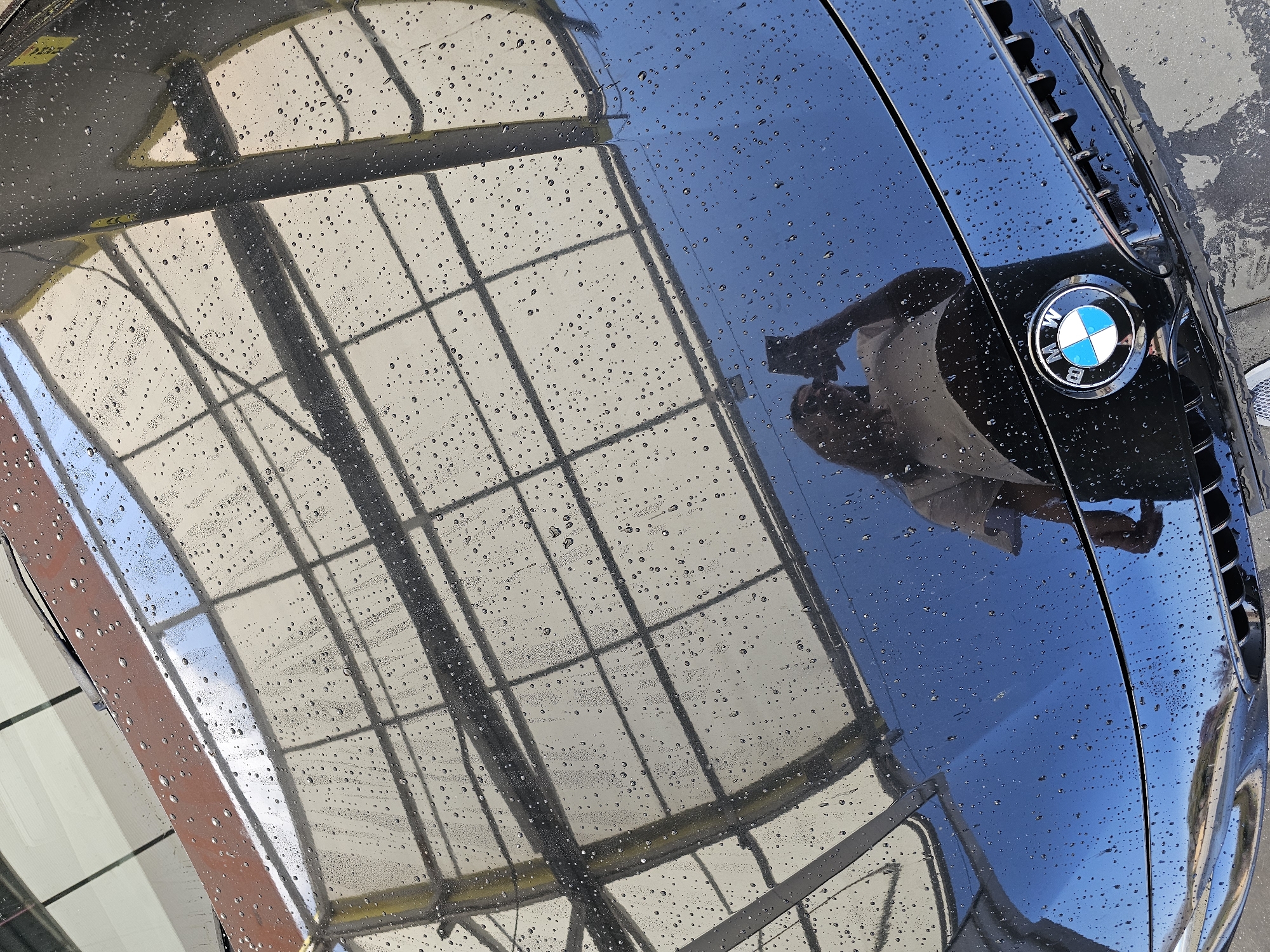 The image depicts the hood of a black BMW car, possibly a newer model, with water droplets on its surface. The BMW logo is visible on the right side of the hood, near the grill. The reflection on the hood shows a metal structure, possibly a car wash or a covered parking area, and also reflects a person holding a phone, presumably taking the picture. It is not possible to determine the exact location in Plovdiv, Bulgaria, based on this image alone.