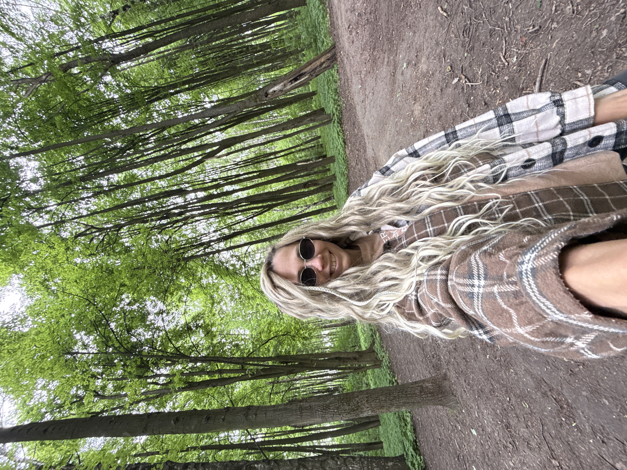 A woman with long, blonde hair and sunglasses is posing on a dirt path in a forest. She is wearing a plaid shirt. The path is bordered by trees with green leaves. The location is Rusanovo, Russia.