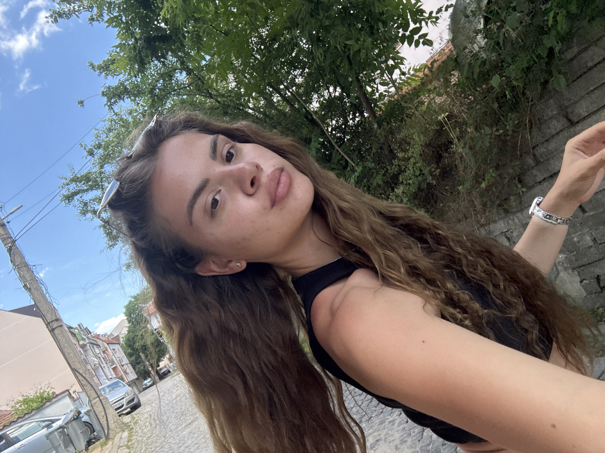A woman is standing outside in Unknown, Unknown, taking a selfie. She has long, wavy brown hair, fair skin, and is wearing a black top. Sunglasses are perched on her head, and she has a silver watch on her wrist. Behind her, the scene is a blend of urban and natural elements. There's a stone wall with greenery growing over it, as well as trees. In the background, there's a street with buildings and cars visible under a blue sky with some clouds. A utility pole is also visible in the background. The ground appears to be paved with stone.