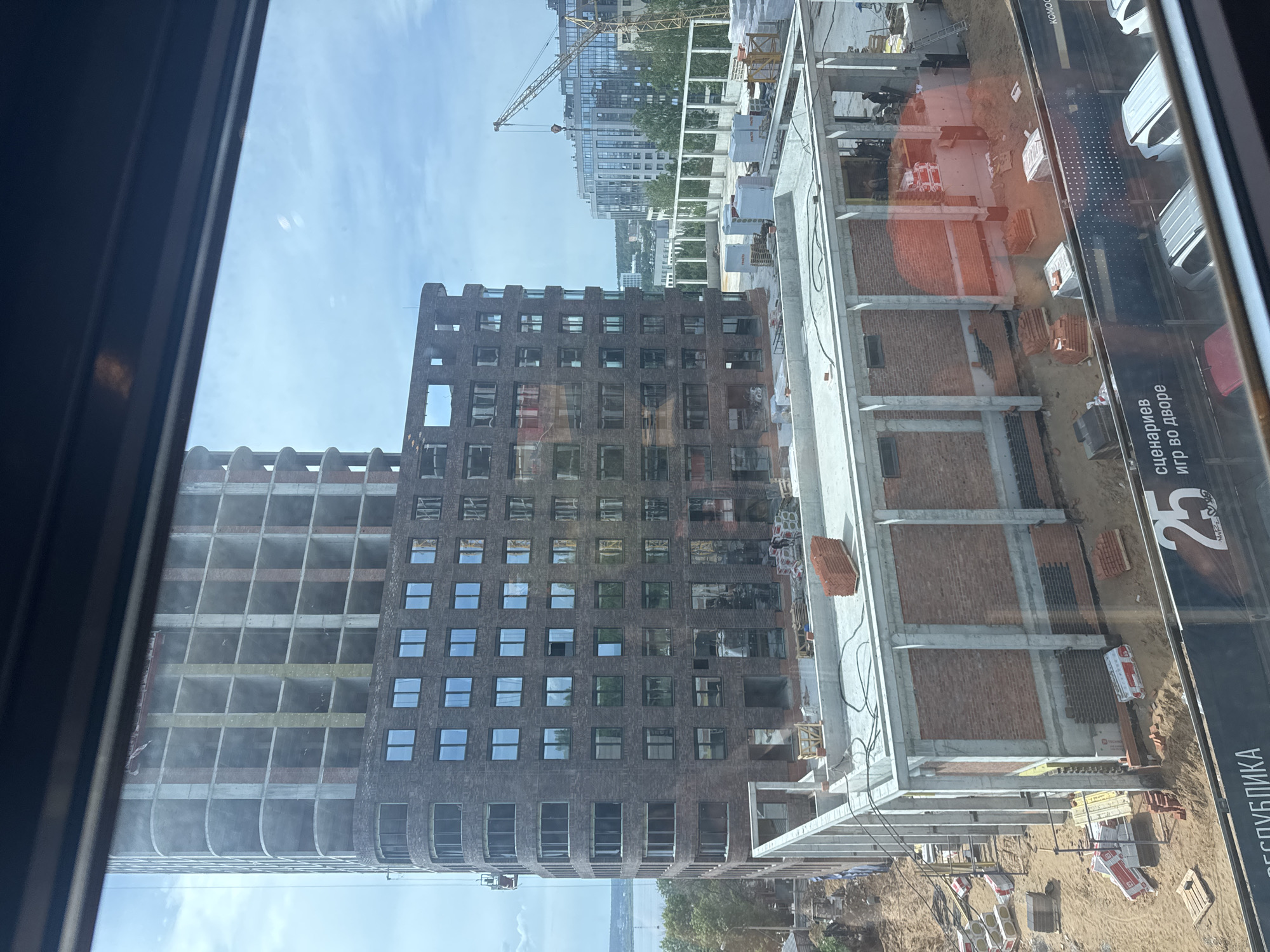 View from a high-rise window onto a construction site in Izhevsk, Russia. Two multistory buildings are under construction: one is a bare concrete frame, and the other has a brick facade partially completed. Construction materials like bricks and scaffolding are visible. A billboard is adjacent to the construction. In the distance, more city buildings and a construction crane can be seen.