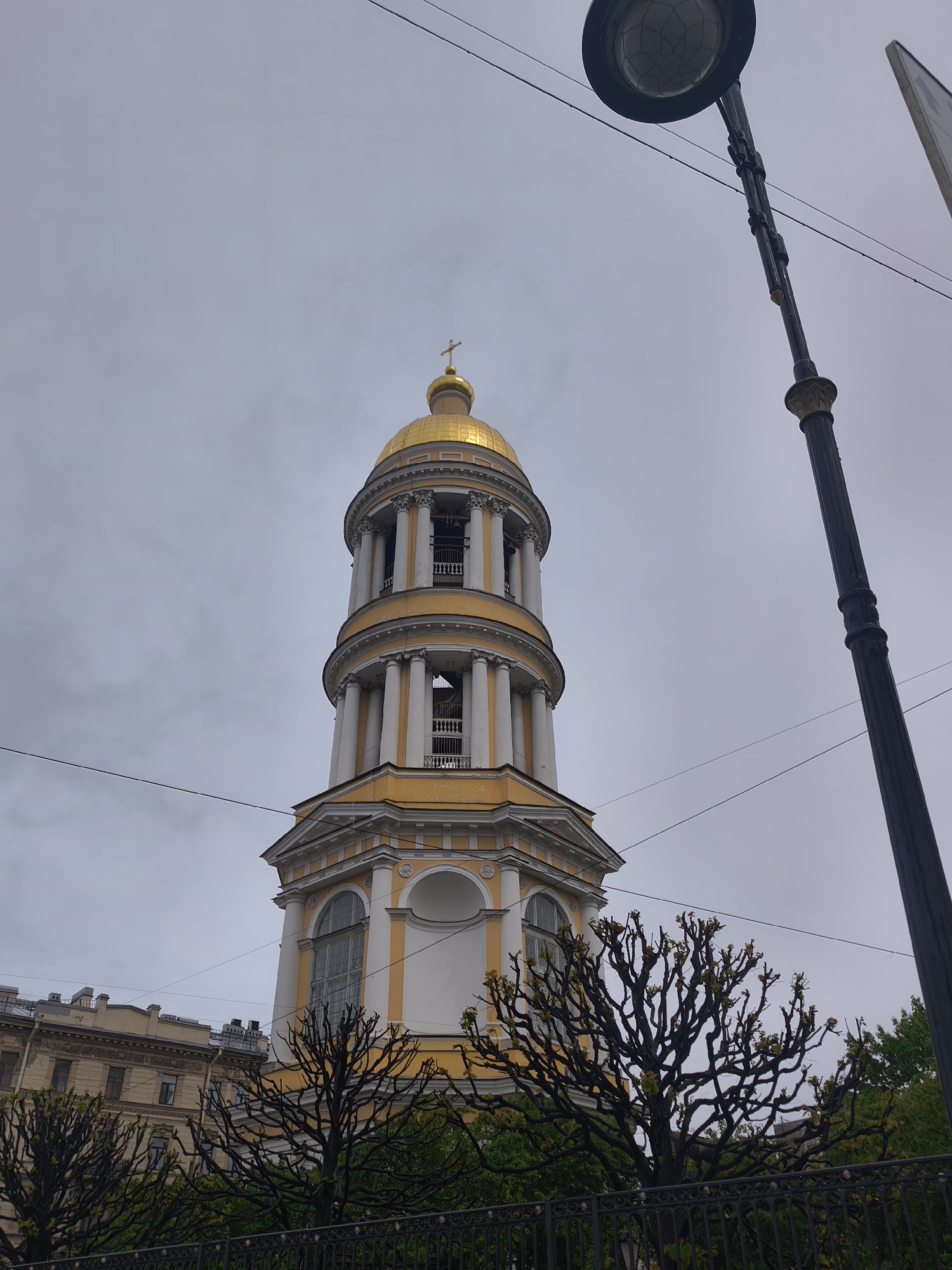 The image shows a tall, ornate bell tower with a golden dome, topped with a cross, rising above trees and buildings. The tower is white and yellow, with columns and architectural details. Power lines stretch across the sky. A street lamp is visible on the right side of the frame. The sky is overcast and gray. The location appears to be an urban area.
