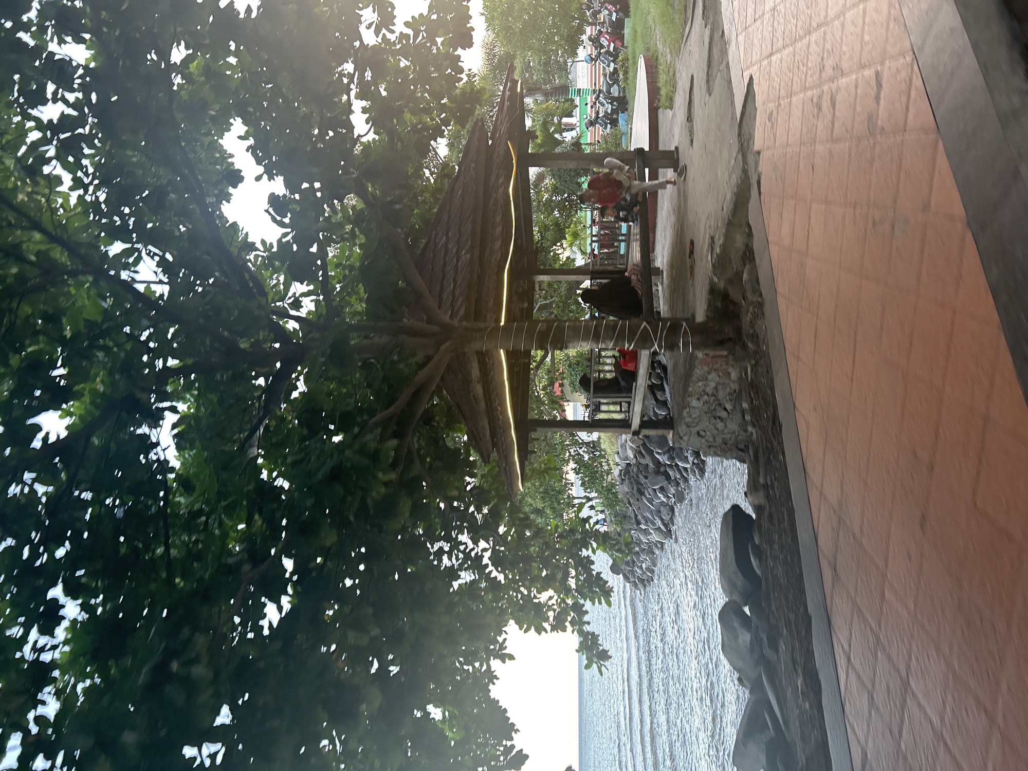 The image depicts a serene waterfront scene at Pantai Seruni, a popular tourist spot in Bantaeng, Indonesia. The foreground is dominated by a paved area with reddish-orange tiles, suggesting a walkway or seating area. To the left, the calm blue sea stretches towards the horizon, meeting a rocky shoreline.
A prominent feature of the scene is a rustic wooden gazebo situated along the coast, providing shade and a resting place for visitors. The gazebo is decorated with string lights, indicating it may be illuminated at night. Several individuals are visible inside and around the gazebo, with some seated and others standing. They appear to be enjoying the view or the company.
Behind the gazebo, a row of motorcycles is parked, suggesting that visitors typically arrive by private vehicle. Lush green trees and vegetation surround the area, adding to the tranquil atmosphere. The overall impression is one of a relaxed, coastal recreational area.