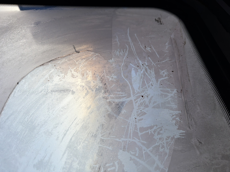 The image presents an interior perspective through a heavily frosted vehicle windshield, observed in the city of Золотая Степь, Russia. The glass is substantially obscured by dense, crystalline frost patterns. A partial, arced area in the upper-center, consistent with a windshield wiper's sweep, exhibits less frost, revealing a hazy exterior background with pale blue and faint orange-pink light, indicative of dawn or dusk. Below this cleared section, the frost features intricate, abstract branching and irregular linear formations, interspersed with scattered dark particulate debris. A small, dark, angular anomaly is visible near the top edge of the frosted area. The lower portion of the image displays the vehicle's black dotted frit band along the windshield's edge, and a faint manufacturer's emblem is discernible in the lower right corner of the glass. The overall scene suggests cold ambient temperatures.