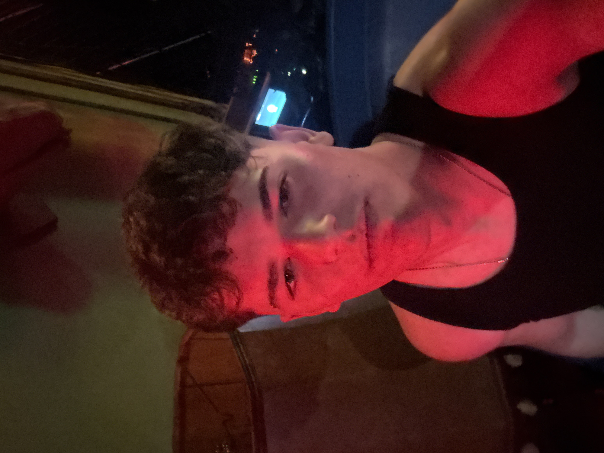 A young person, possibly male, with curly brown hair and wearing a black top, is captured in a dimly lit, indoor setting under dominant red artificial lighting. A thin silver chain necklace is visible around their neck. The person's face, partially obscured by shadows and highlights from the intense red light, is angled towards the viewer, displaying a neutral to slightly pensive expression. Their skin and clothing are bathed in a strong red hue. In the background, a distinct bright blue light source, possibly a phone screen or a digital display, is visible to the right of the person's head, contrasting sharply with the pervasive red. Other faint, indistinct lights and dark elements suggest a complex, atmospheric background, potentially in a bar or club. Parts of what appear to be wooden furniture or fixtures are visible in the lower corners. The image is taken from an oblique angle, lending a dynamic perspective. No text is visible in the frame. The scene evokes a nighttime social event in Sofia, Bulgaria.