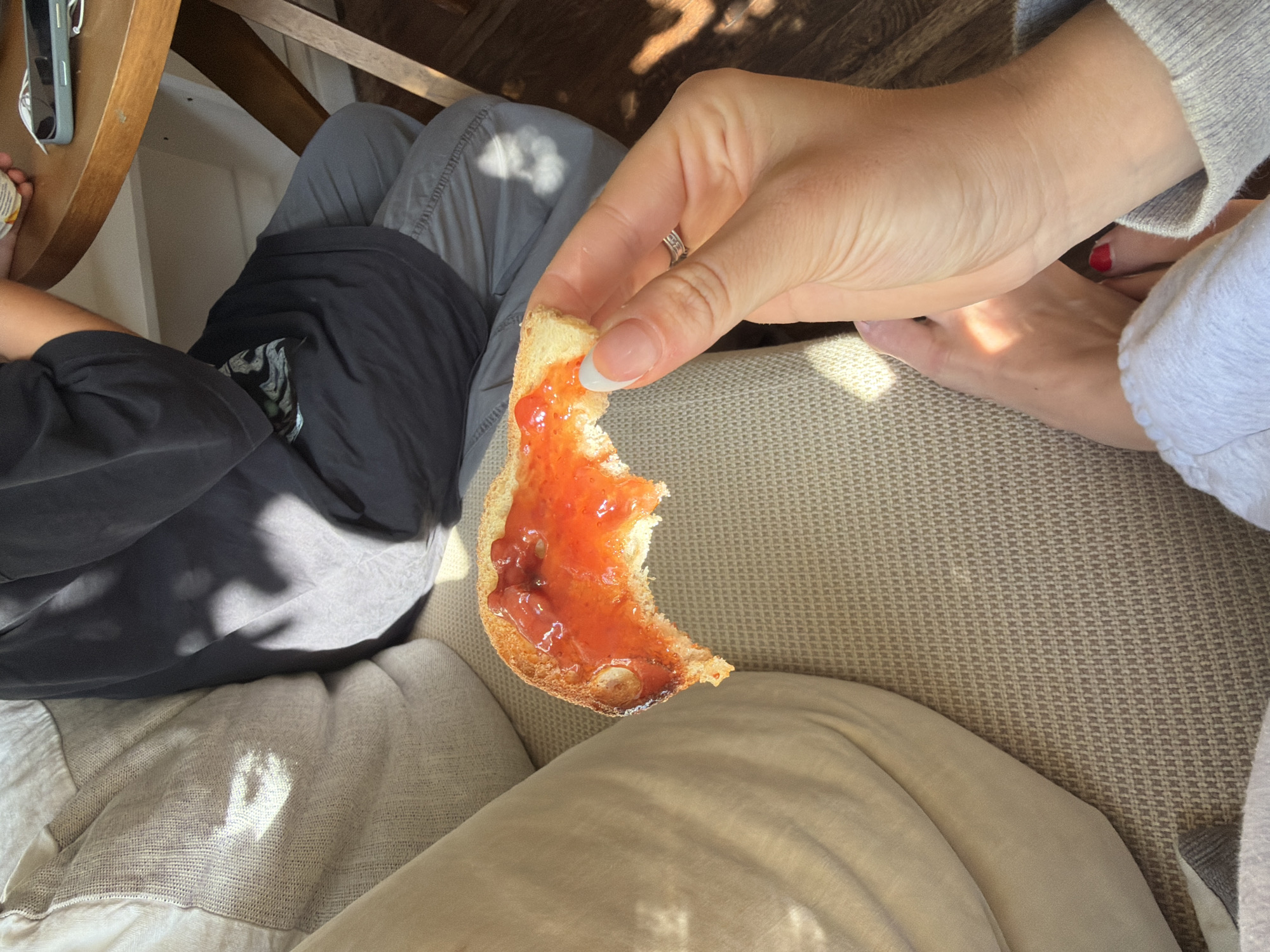 The image captures a relaxed, casual moment indoors, likely in Los Angeles, USA, bathed in bright natural sunlight. The central focus is a close-up of a person's right hand holding a partially eaten piece of toast generously spread with glossy, bright red jam or preserves. The toast has a slightly burnt edge, indicating it was toasted. The hand displays a neat French manicure with white tips and a silver ring on the ring finger.
To the left, a second person is partially visible, seen from above. They wear a dark short-sleeved shirt and light grey pants. Their left arm rests on a round wooden table, where a grey-blue smartphone with white wired earbuds connected to it is placed.
Below the hand holding the toast, light beige, textured upholstery suggests a comfortable sofa or armchair where the first person is seated. Further down, the first person's bare feet are visible, with one foot showing vibrant red painted toenails, resting on a light-colored blanket or fabric. The second person's legs are also covered by light-colored fabric, possibly their pants or a blanket.
Strong, dappled sunlight streams into the scene, creating distinct shadows on the wooden table, the fabrics, and the floor, indicating a sunny daytime setting, possibly morning or late afternoon. The overall atmosphere is one of leisure and comfort, capturing a simple moment of enjoying a snack. No visible text is present in the image.