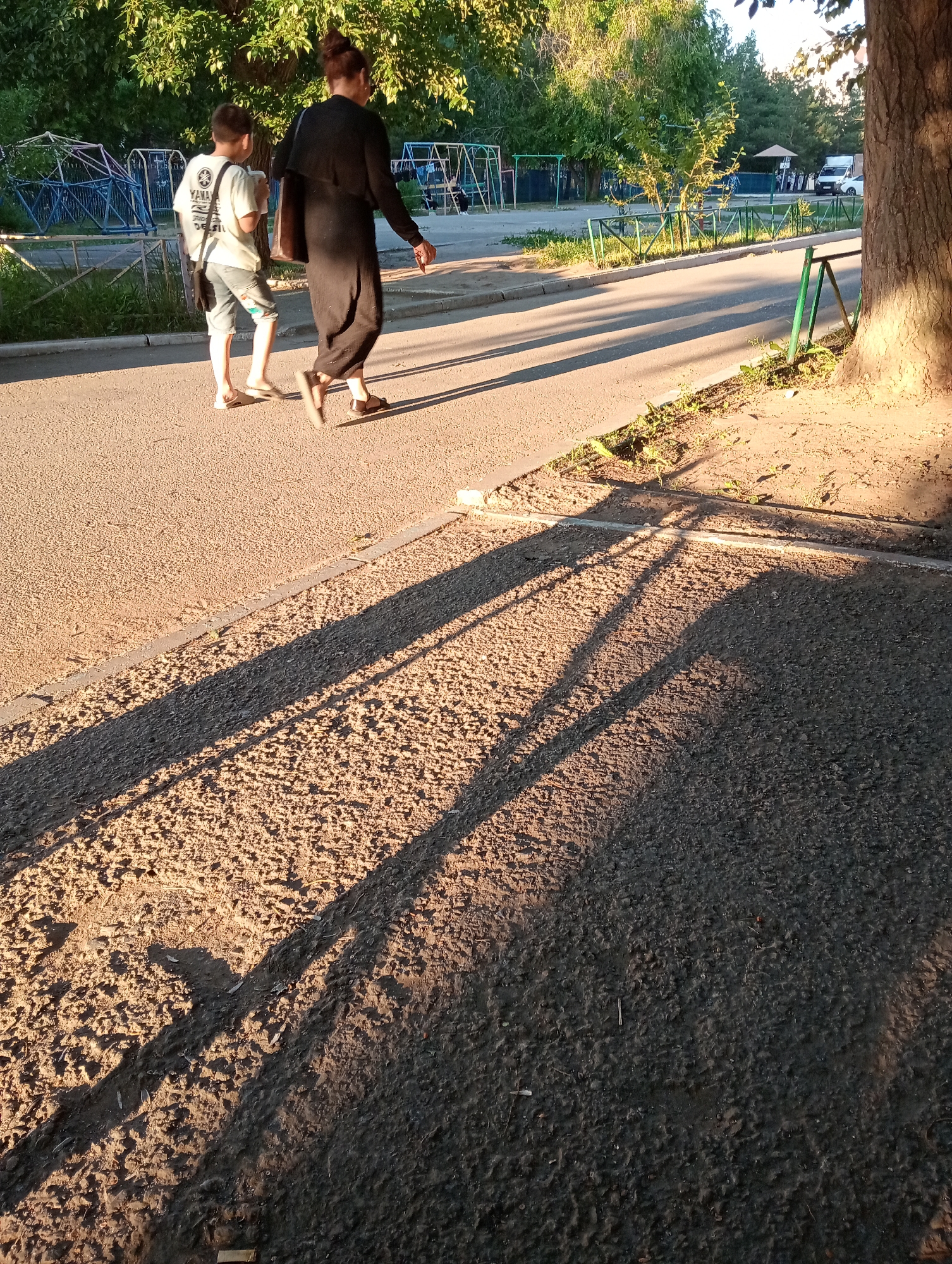 Two individuals, an adult and a child, walk on a paved path bordered by grass and a fence. The adult, wearing a black dress and carrying a bag, walks ahead of the child, who is wearing a white t-shirt and shorts and holding a cup. Sunlight casts long shadows on the path. In the background, playground equipment and trees are visible. The scene appears to be set in a park or recreational area within an urban environment.