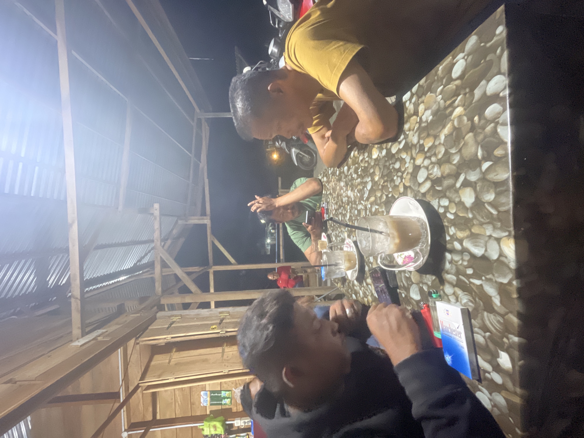 Four men are seated at a table under a corrugated metal roof supported by wooden beams. The table is covered with a cloth depicting pebbles. In the foreground, a man in a black hoodie is looking down at his phone. To his right, a cigarette pack and two lighters are on the table. A glass of iced coffee sits in front of him. To the right of the table, a man in a yellow shirt is looking away from the camera, with his arms crossed. Behind him, another man in a green shirt is looking at his phone, with his hand near his forehead. Further back, a man in a red shirt is visible. Motorcycles are parked in the background. The scene appears to be an outdoor cafe or rest stop in Cotkruet, Indonesia, possibly at night given the lighting conditions and lack of natural light.