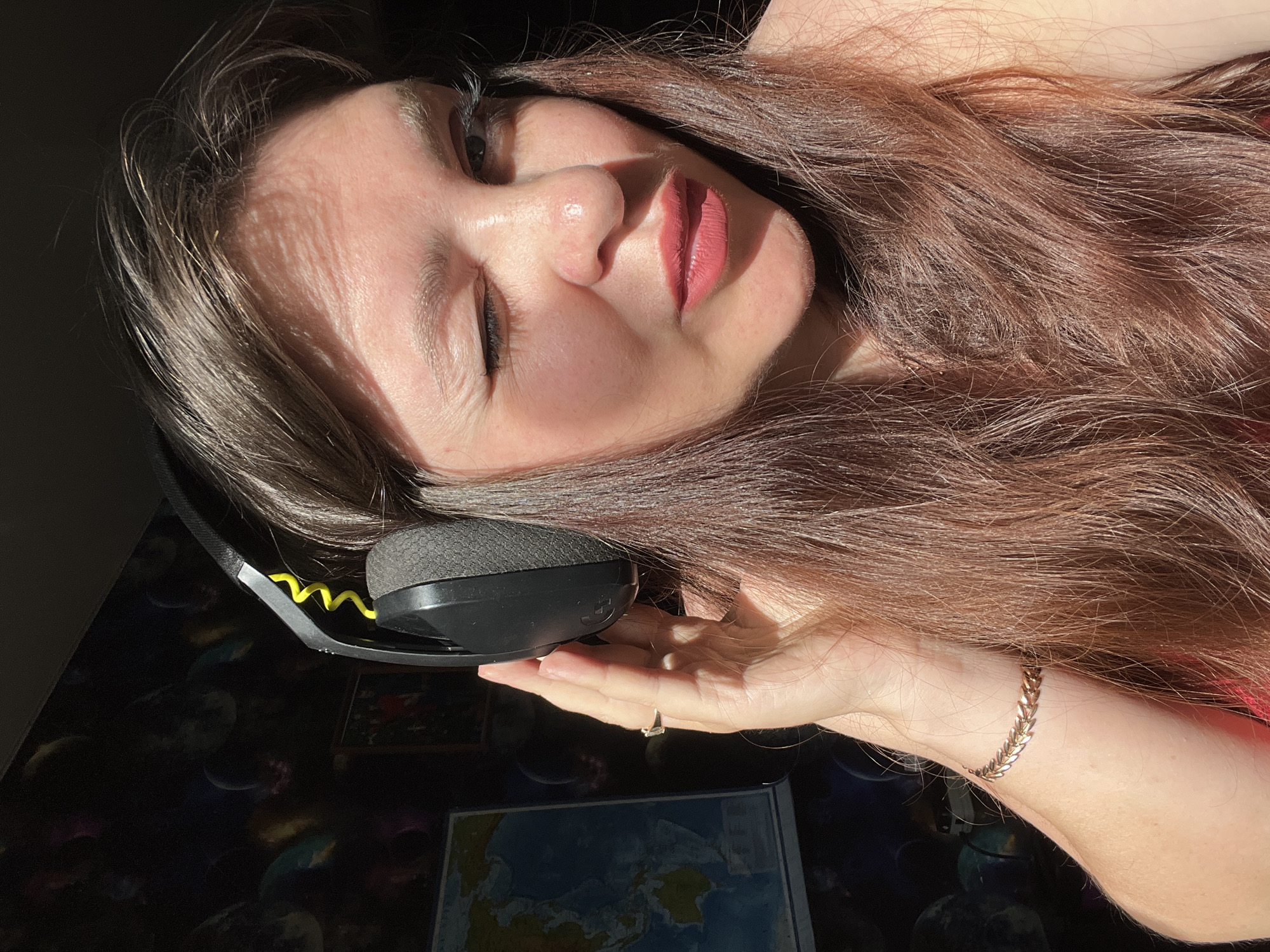 A woman with long brown hair wearing headphones is captured in a close-up, outdoor shot. She is winking and her right hand is holding the headphone on her ear. She is wearing a pink top and a gold bracelet. The background shows a wall with a planetary-themed wallpaper and a map. The lighting suggests it is daytime, with sunlight creating shadows on her face. There is no indication of the specific location in Noviy, Russia.