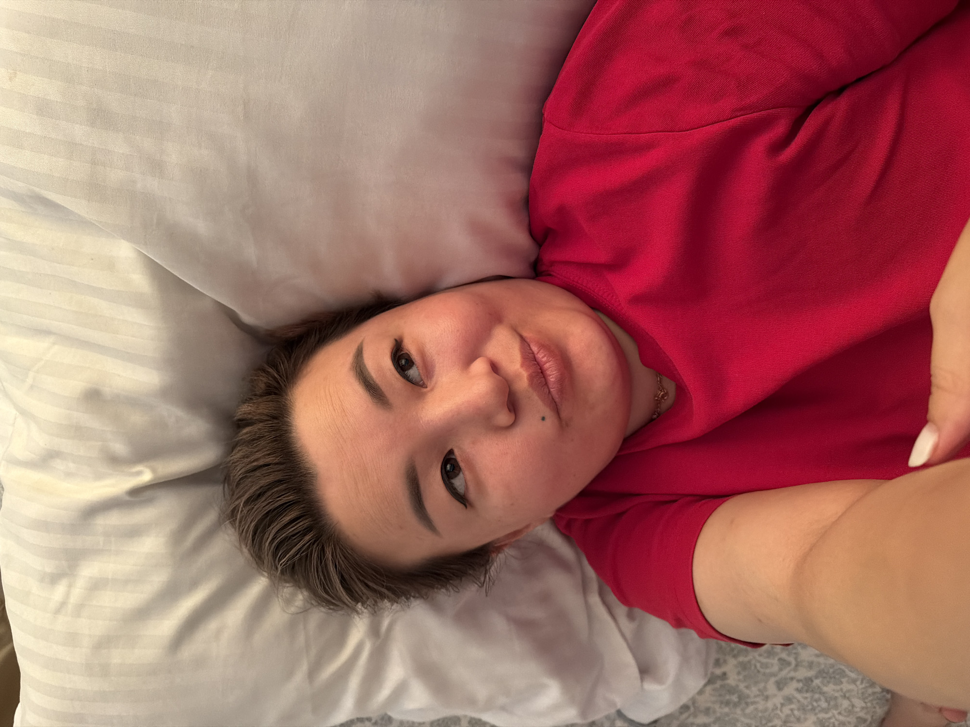 A young woman is lying in bed, propped up on a pillow. She is wearing a red t-shirt. Her hair is short and brown. She is looking up and to her right with a slight pout. The background consists of a white pillow and a duvet cover with a gray and white pattern. The image appears to be a selfie taken from a slightly high angle. There is no indication of the specific location in Astana, Kazakhstan.