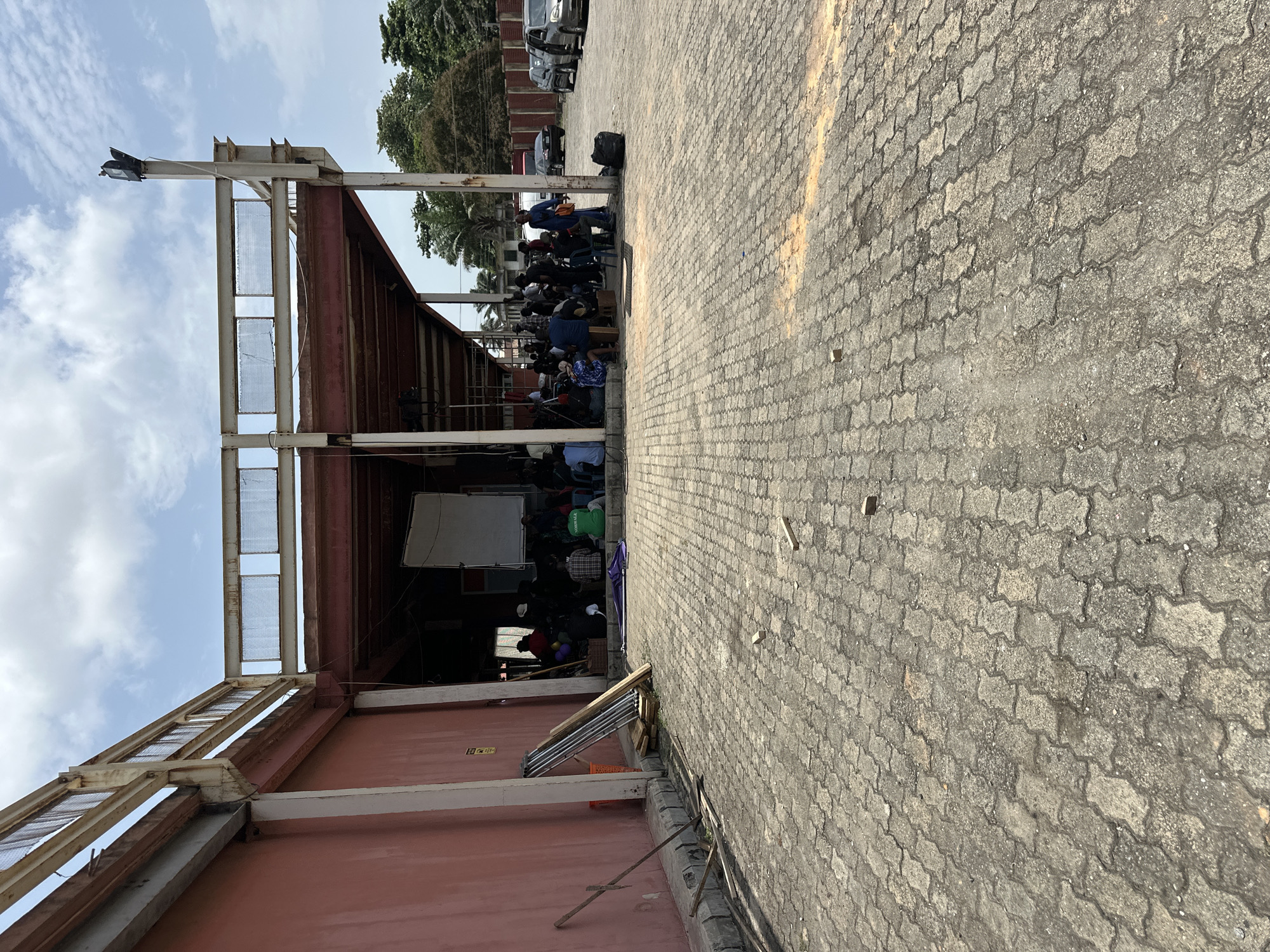 The image depicts an outdoor gathering in Lagos, Nigeria. People are congregated under a metal structure with a roof. Cars are parked in the background. The foreground is a paved area. There is a whiteboard visible within the structure. Ladders are leaning against the structure. The sky is visible in the upper portion of the frame.