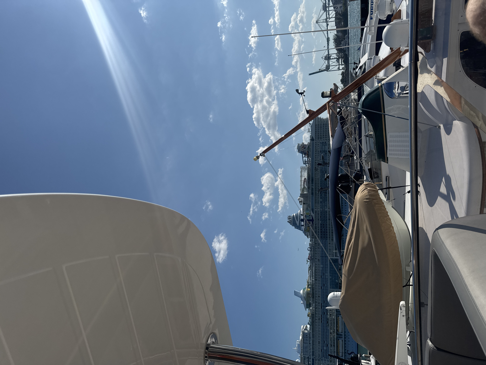 The image shows a view from a boat in Miami Beach, Florida, United States of America. The sky is blue with some clouds. The camera looks out toward the port of Miami, where large cruise ships are docked. The foreground includes parts of the boat, such as the white canopy and railing, as well as a covered dinghy. The cranes from the port and some other nearby boats are also in the frame.