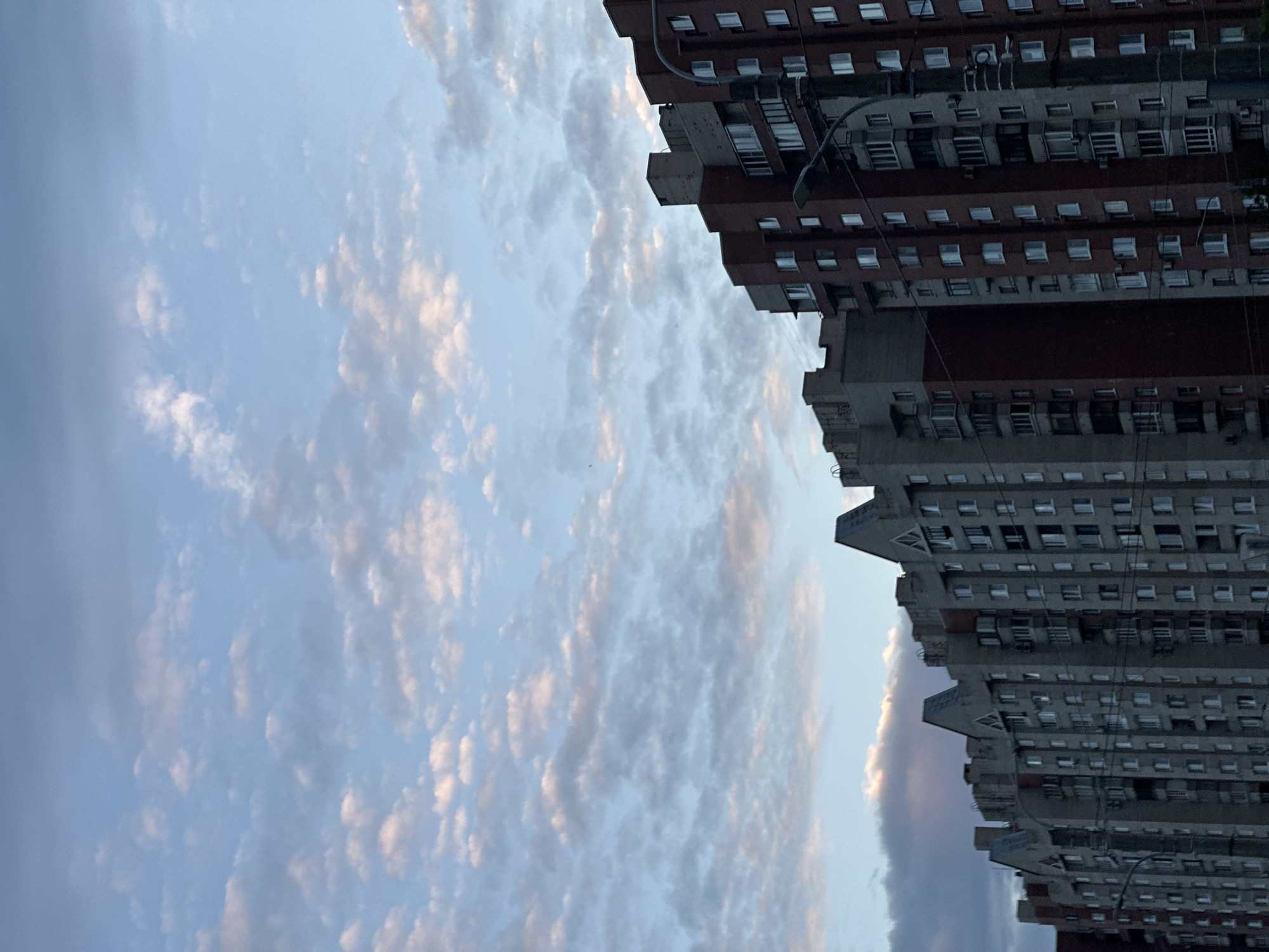 The image shows a street view in Saint Petersburg, Russia, looking up at apartment buildings against a cloudy sky. The buildings are tall and appear to be made of concrete and brick, with numerous windows and balconies. The sky is a light blue with scattered white and pinkish clouds. The overall impression is of an urban residential area during twilight.