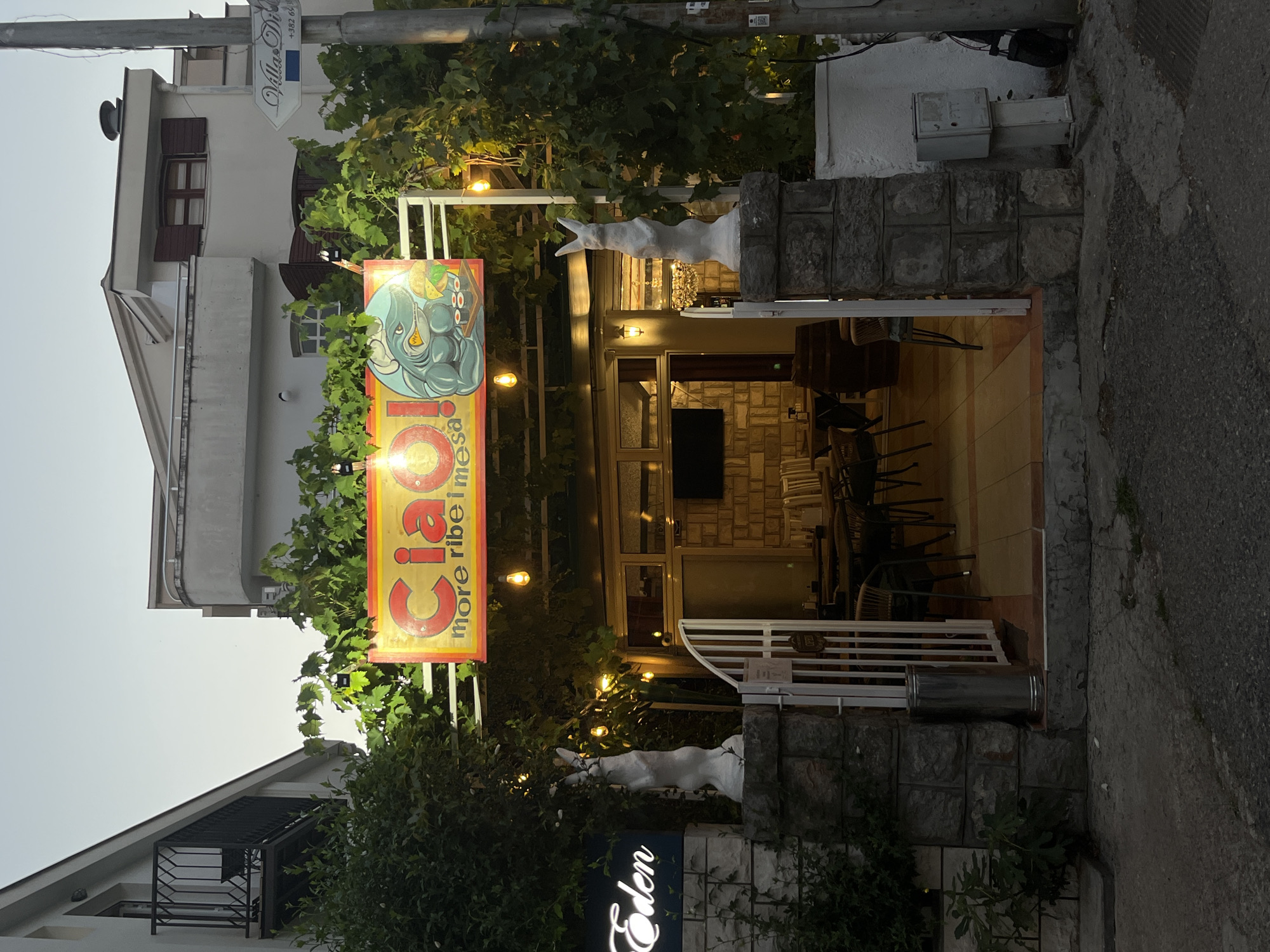 A horizontal, eye-level outdoor shot captures the entrance to a restaurant in Budva, Montenegro, as dusk settles. The restaurant features a lively sign that reads "Ciao! more ribe i mesa." Beneath the sign, a white archway opens into a seating area with stone walls, tables, and chairs. Vines drape over the archway, interspersed with small, glowing lights. To the left of the entrance, a neon sign displays the word "Eden" in white. Two white rabbit statues stand guard on stone pillars flanking the entrance. A tall, cylindrical silver trash can is positioned to the left of the gate. In the background, apartment buildings with balconies and shutters rise. To the right of the entrance, a street sign points towards "Villas Di..." and shows a phone number. The ground in front of the entrance is paved with asphalt and cracked concrete.