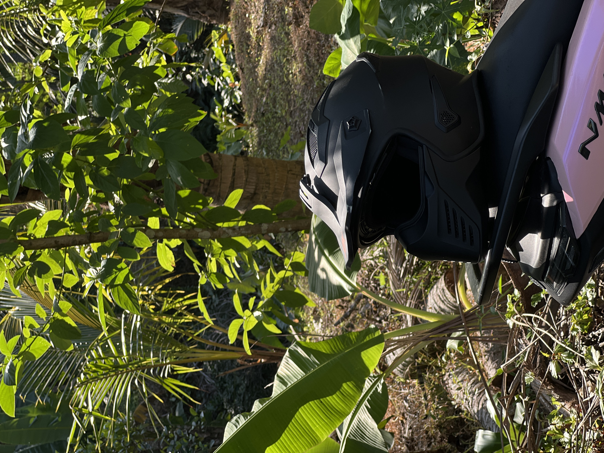 A black motocross helmet rests on the pink seat of a motorcycle, positioned in a lush, tropical outdoor environment. The helmet is matte black with a dark visor and intricate detailing, including vents and a chin guard. Behind the motorcycle, vibrant green palm fronds and banana leaves are visible, illuminated by dappled sunlight. In the background, the scene transitions to a gentle slope covered in dry grass and scattered palm tree trunks, suggesting a rural or semi-rural setting in Kerambitan, Indonesia. The overall impression is one of vibrant natural beauty contrasted with the presence of modern recreational equipment.