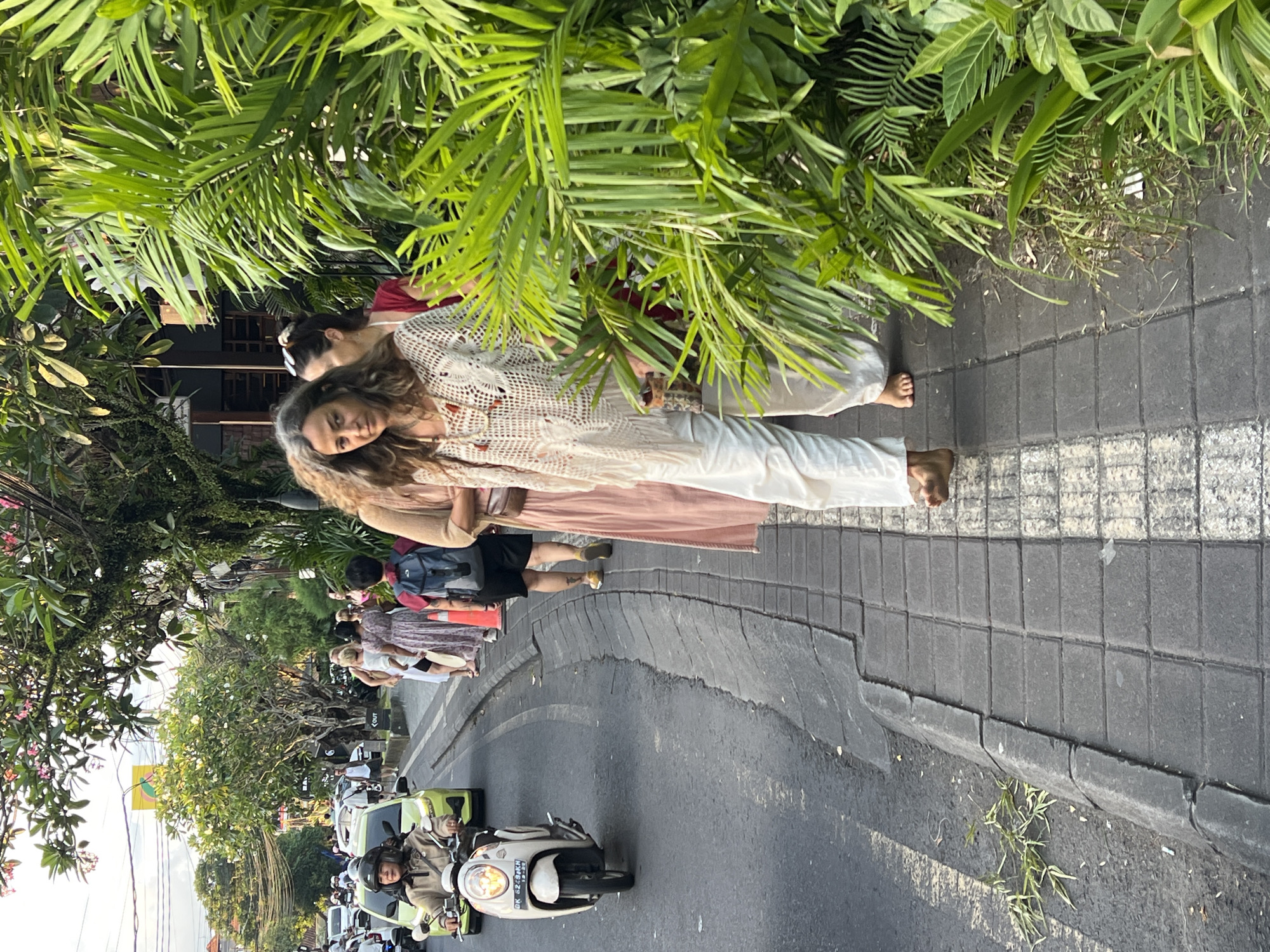 The image captures a vibrant street scene in the Ubud District of Indonesia. A woman with long, wavy hair stands barefoot on a tiled sidewalk, partially obscured by lush green foliage. She is wearing light-colored clothing. Another person stands near her, also barefoot. A line of people extends along the sidewalk behind them. The street is filled with scooters and cars, indicative of traffic. Trees and tropical plants are abundant, contributing to the area's natural atmosphere.