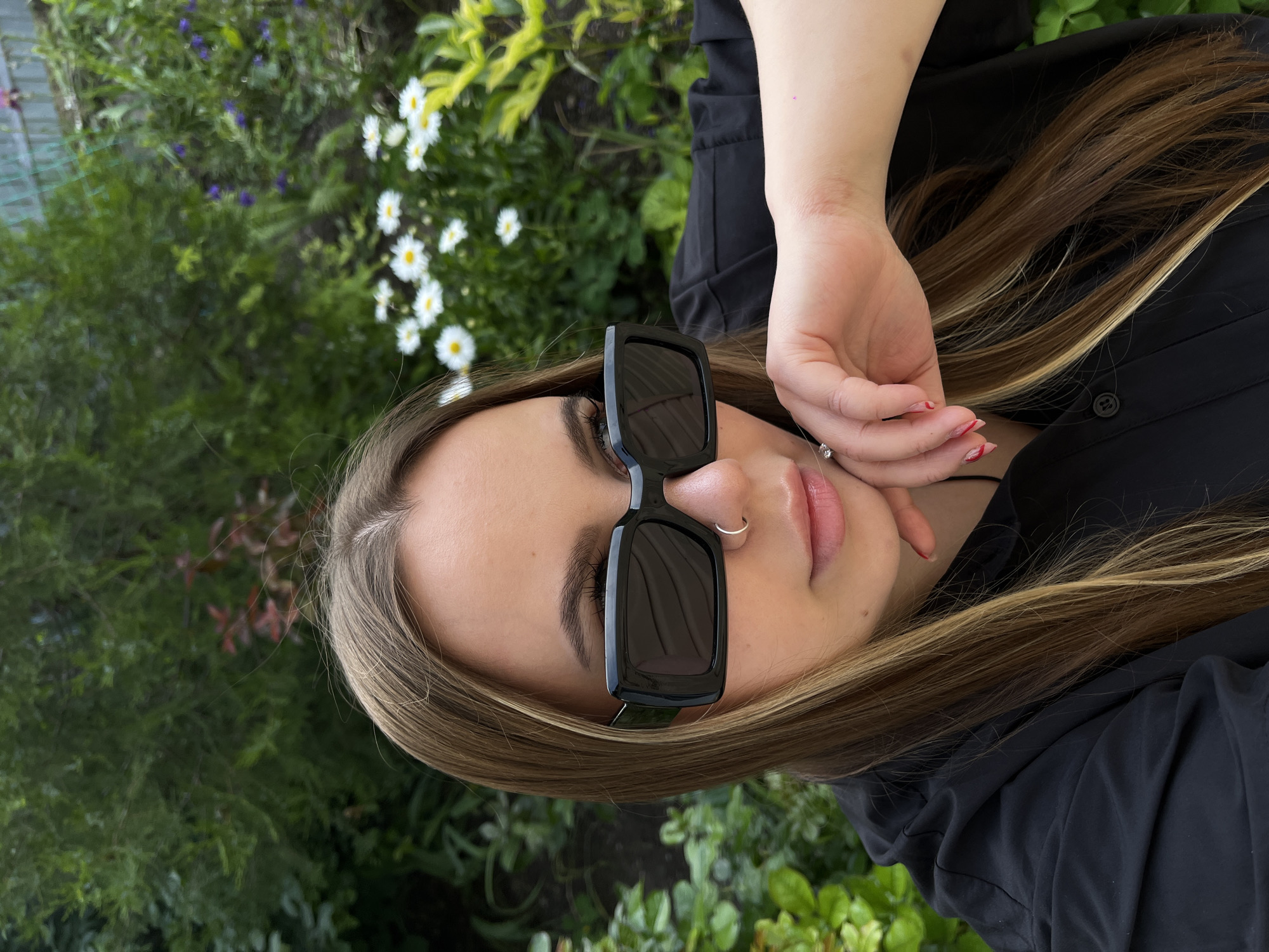 A close-up, rotated portrait shows a young woman posing outdoors in Novopokrovskaya, Russia. Her head and upper torso are featured prominently against a softly blurred background of abundant green foliage interspersed with small white daisy-like flowers and a few purple blooms.
The woman has long, straight hair with a mix of brown and blonde highlights, framing her face and resting over her black collared shirt. She is wearing large, black rectangular-framed sunglasses that conceal her eyes. A small, silver hoop nose ring is visible on her right nostril. Her lips are slightly parted, showcasing a natural pink shade. Her right hand is raised, with fingers gently positioned near her chin and jawline. Her fingernails are painted with a vibrant red polish, which shows minor chipping on some digits. A delicate silver pendant hangs from a thin chain, visible near her neck. The natural lighting suggests it is daytime. No text is visible in the image.