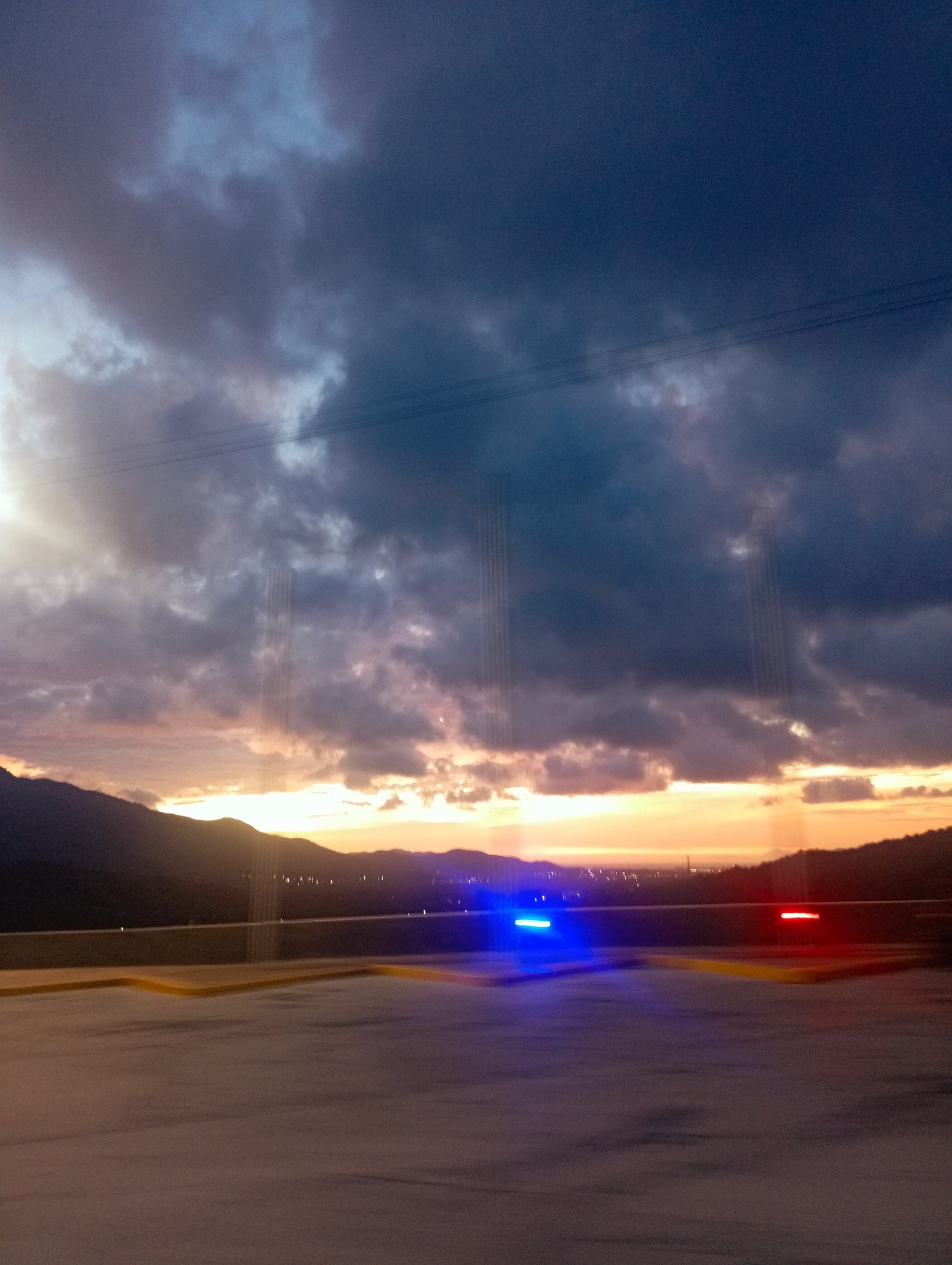 The image depicts a view from what appears to be a parking garage or elevated platform in Altagracia, Venezuela. In the foreground, there is a section of gray concrete surface with yellow safety barriers, with a horizontal streak of blurred blue light on the ground. In the middle ground, there is a mountain range silhouetted against a sunset or sunrise, where the sky is a mix of orange, pink, and purple hues. The background is dominated by dark, dramatic clouds. Visible between the mountains are scattered lights, possibly from the city. A faint power line crosses the top of the image. The overall impression is of a landscape viewed from a higher elevation point within an urban environment.
