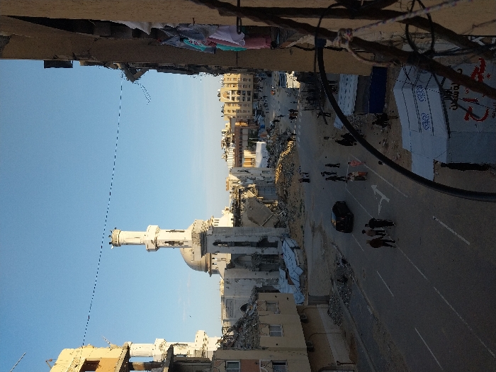 An elevated view captures a street scene in Khan Yunis, Palestinian Territory, characterized by extensive structural damage to buildings under a clear blue sky. In the mid-ground, a mosque with a prominent central dome and two minarets stands partially damaged; significant portions of its surrounding structures are reduced to rubble. One minaret appears largely intact, while the other shows damage. To the left and right of the mosque, multi-story buildings exhibit severe destruction, with facades collapsed, exposing interiors and leaving large piles of concrete and twisted metal debris on the ground.
On the asphalt street below, a single dark-colored car is parked or slowly moving. Numerous pedestrians, individuals and small groups, are present on the street and sidewalks, navigating around the debris. White road markings, including a double solid line and a right-turn arrow, are visible on the road surface.
In the immediate lower right foreground, partially obscured by a metal railing and wires, a white tent-like structure with Arabic script is erected. Laundry, including shirts and other garments, hangs from a structure on the far right, suggesting a habitable building adjacent to the vantage point. Buildings in the distant background appear less damaged, forming a relatively intact urban skyline.