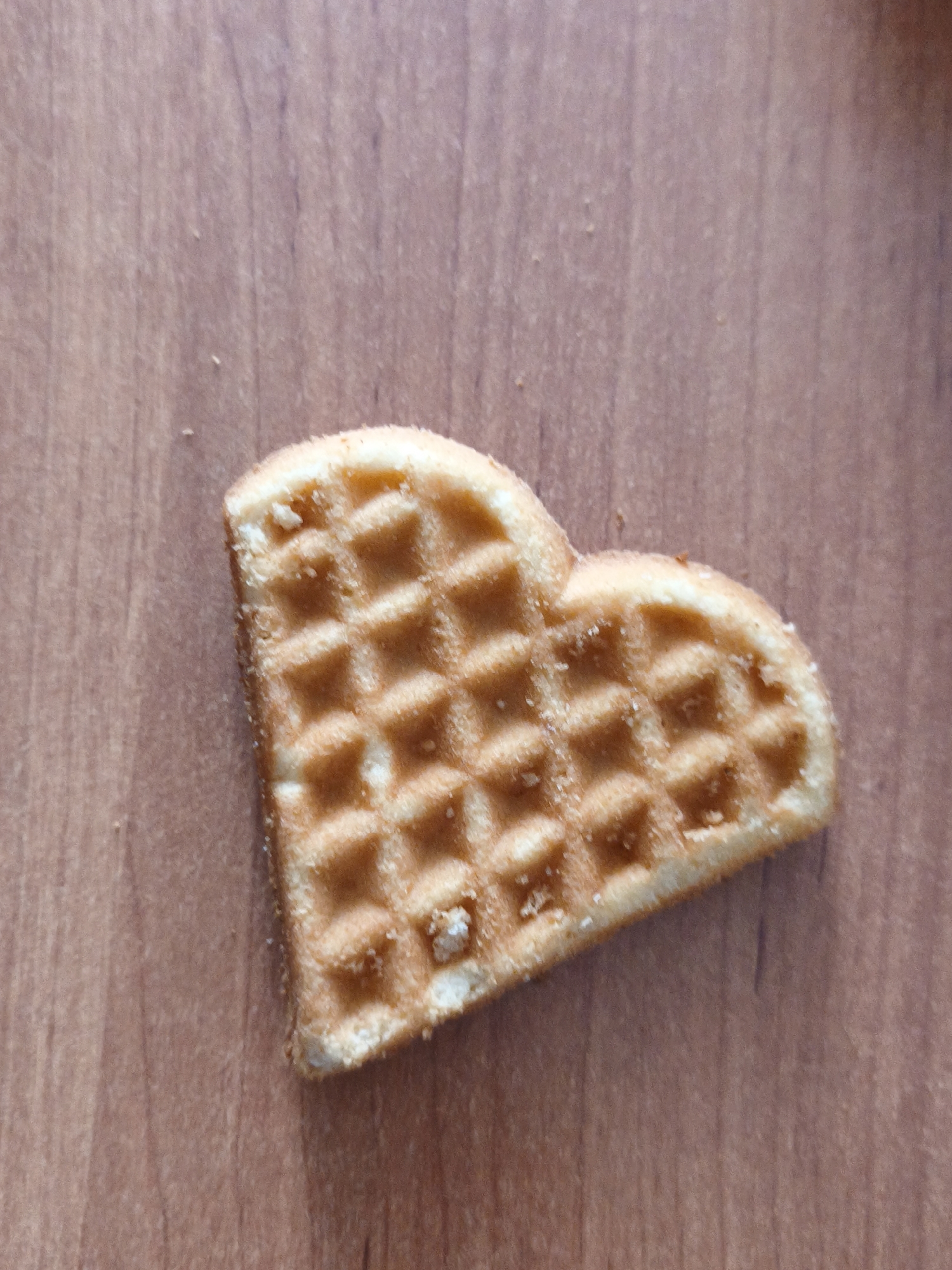 A single heart-shaped waffle sits on a wooden surface. The waffle is golden brown with a textured grid pattern. The surface appears to be a light-colored wood with a visible grain. There are no people or animals in the image. The location is not discernible from the visual information provided.