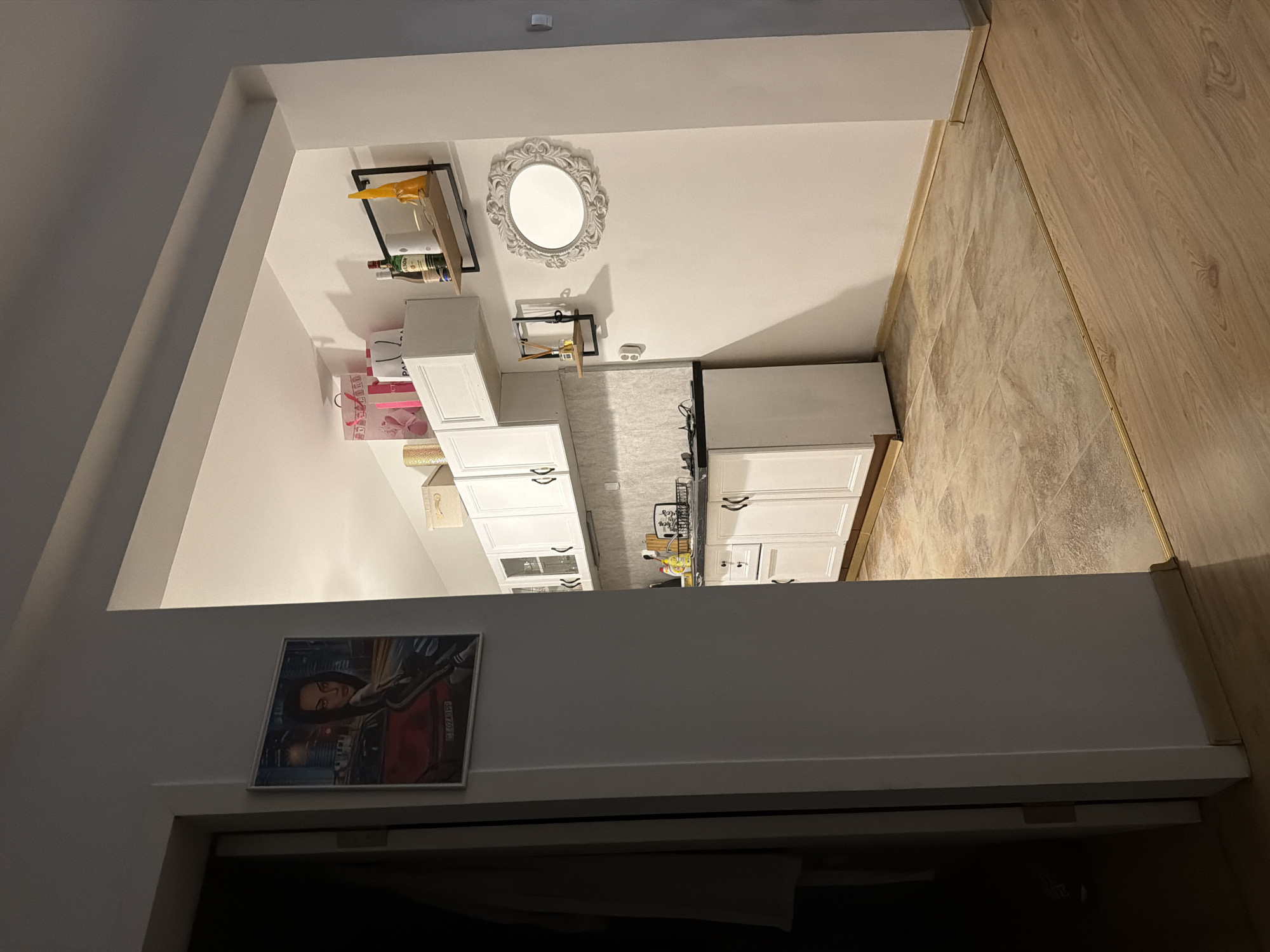 The image depicts the interior of a kitchen and an adjacent hallway, possibly in a residential building in Plovdiv, Bulgaria. In the foreground, a doorway reveals a hallway with a partially visible door on the left and a white wall on the right. The floor in the hallway transitions from a light-colored wood laminate to a beige tiled floor.
The kitchen area is visible through an archway. It features white upper and lower cabinets. On the wall above the lower cabinets, there are several items including bottles of alcohol, a decorative mirror with an ornate frame, and two small shelves holding decorative objects. The upper cabinets are topped with several gift bags. The countertop is dark, and a sink with a faucet is visible. The floor in the kitchen is tiled with a beige and brown patterned tile.
On the wall in the hallway, to the left of the archway into the kitchen, a framed picture of a woman leaning against a red car is displayed. The image is captured from a medium distance, providing a clear view of the kitchen's layout and some of its contents. The lighting suggests it is indoors, possibly at night or in a dimly lit environment. There are no people visible in the image.