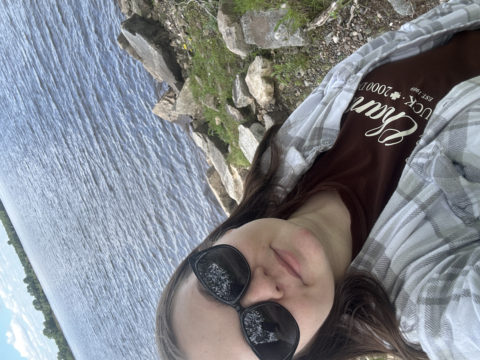 The image, when correctly oriented, depicts a person relaxing by a large body of water with a rocky and grassy shore. The subject, a person with long dark hair, is captured from the chest up, wearing dark sunglasses that reflect trees. Their expression is relaxed, with a slight smile and partially closed eyes behind the sunglasses. They are dressed in a brown t-shirt layered under a light grey and white plaid button-up shirt, worn unbuttoned.
Visible text on the brown t-shirt includes the prominent word "Charm" in a large, elegant script font. Above and around it, smaller text reads "LUCKY", accompanied by a four-leaf clover icon, "2000D", and "EST. 1989".
The background features a vast body of water, likely the Kama River or a reservoir, characteristic of the Chaikovsky, Russia, region. The water's surface shows subtle ripples. Along the right side, a rugged, rocky bank with patches of green grass is visible. In the distance on the left, a treeline marks the opposite shore beneath a bright, partly cloudy sky. The scene evokes a tranquil, daytime outdoor setting.