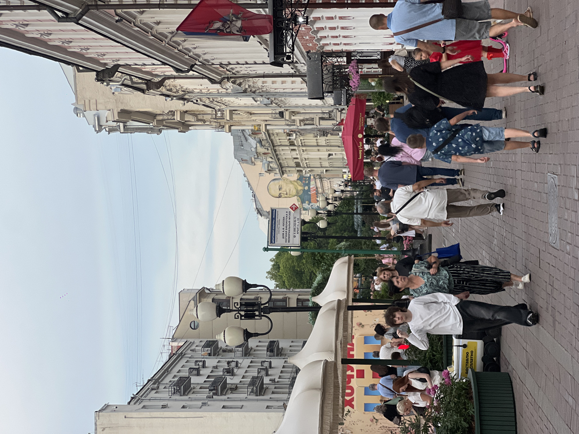 The image shows a street scene in Moscow, Russia, on a slightly overcast day. The street is lined with multi-story buildings, typical of European urban architecture, with balconies and decorative facades. A large mural of a man is visible on one of the buildings. The street is paved with brick and is filled with pedestrians walking in both directions. People are dressed in casual summer clothing. There are shops and cafes on the ground floor of the buildings, some with outdoor seating and awnings. Streetlights and greenery are also present. The overall impression is of a lively, bustling urban environment.
