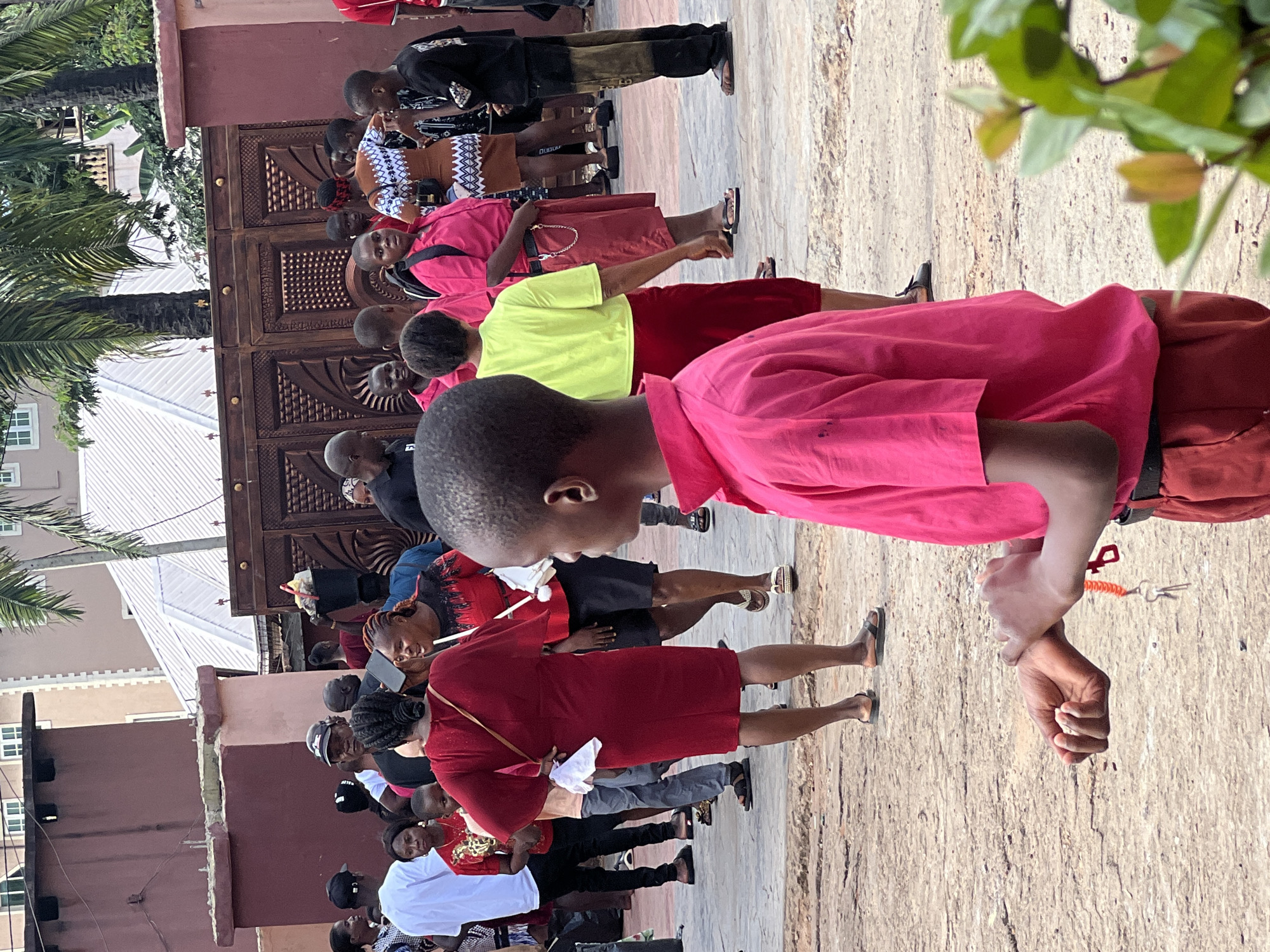 The image shows a street scene in Nnewi, Nigeria, with a group of people gathered outdoors. In the foreground, a young boy in a bright pink shirt and red shorts is facing away from the camera, looking down at his hands. He is holding a set of keys on an orange lanyard. Behind him, a woman in a dark red dress with a light-colored bag slung over her shoulder stands with her back to the camera, her hand on her hip.
Further back, a diverse group of individuals are seen in various casual attire. Some are looking at each other, while others appear to be observing something off-camera. The background features a large, ornate wooden gate, a building with a light-colored roof, and palm trees. The ground is a rough, paved surface. The overall impression is of a community gathering or event taking place on a street in a residential or commercial area of Nnewi.