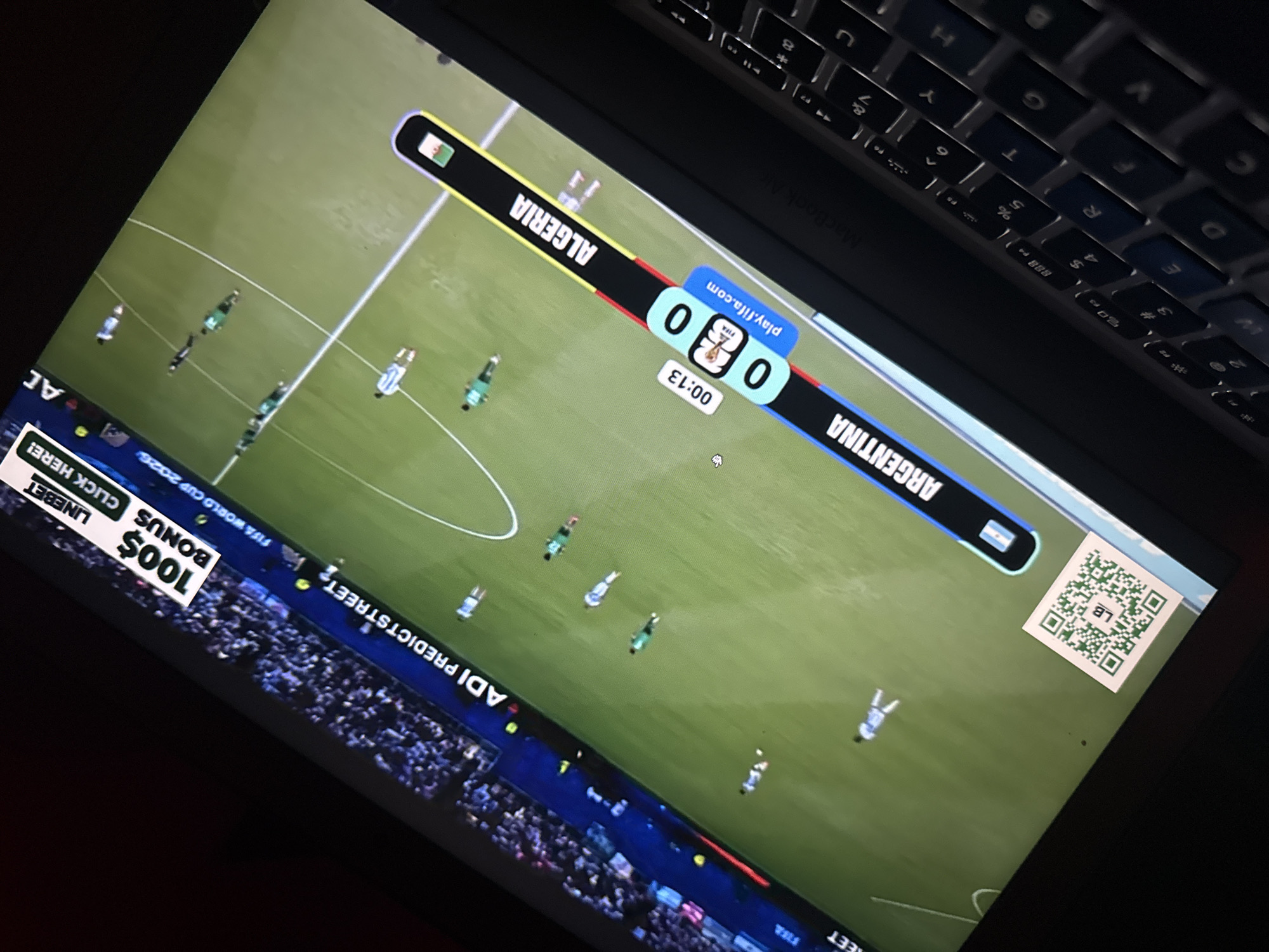 The image shows a laptop displaying a soccer match, likely a video game, with a score overlay. The match appears to be between Argentina and Algeria, with the score currently 0-0 and the time at 00:13. There's a QR code in the bottom left corner. The overall scene is dimly lit. The image is taken from an angled perspective, showing part of the laptop's keyboard. There is no indication of the city of Kufena, Nigeria, in the image.