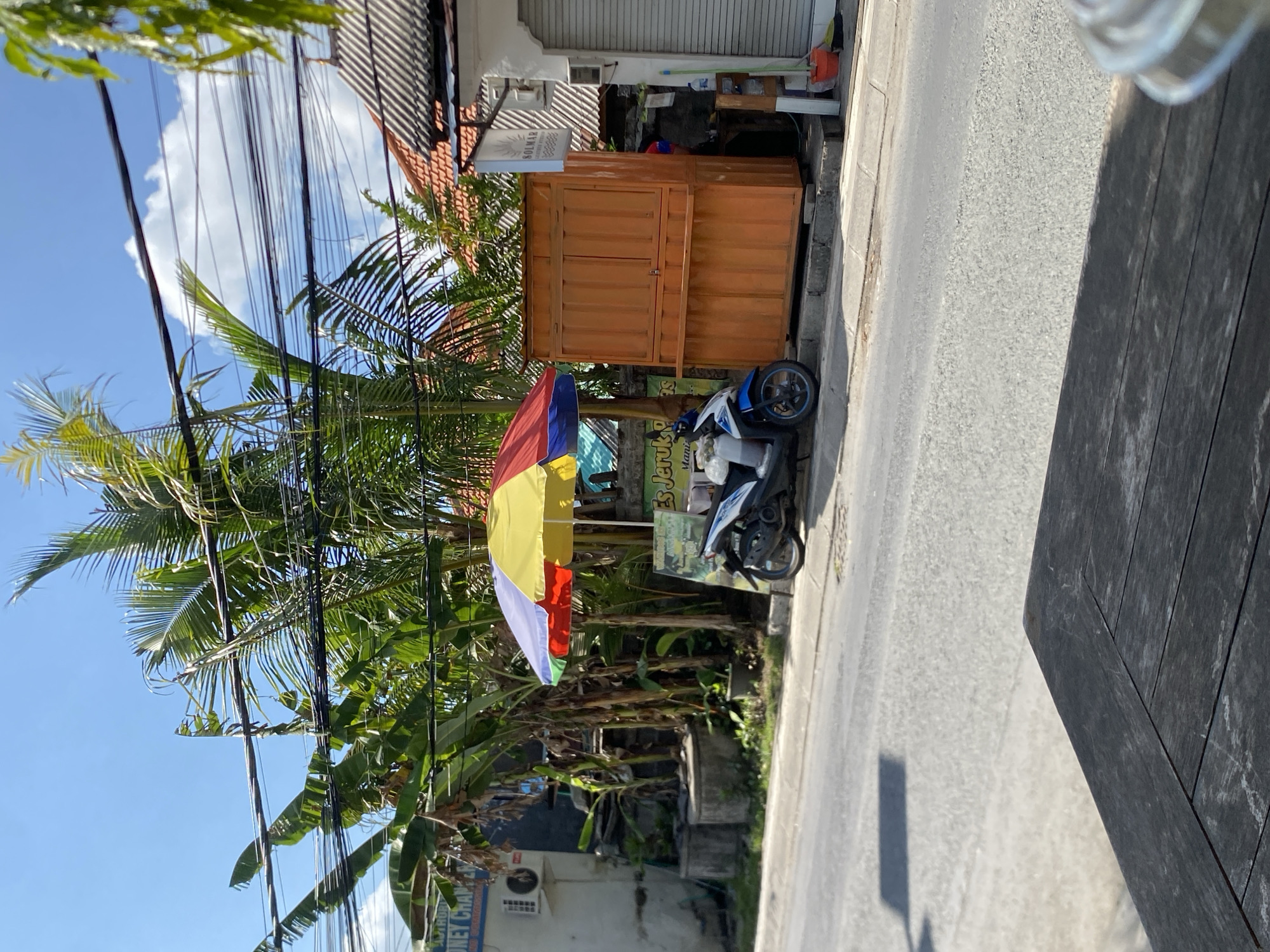 The image depicts a street scene in Mengwi, Indonesia. A blue and white scooter is parked on the side of the road next to an orange wooden kiosk. Above the kiosk, a colorful umbrella, striped with red, yellow, and blue, is open. Behind the kiosk and umbrella, there is lush vegetation, including banana trees and palm trees, with power lines strung above them. To the left of the kiosk, a white building with an air conditioning unit is visible, and a sign for "MONEY CHANGER" is present. The road in front of the scene is paved, and a dark wooden table occupies the foreground, with a portion of a clear cup visible on the right edge. The sky is blue with scattered white clouds.