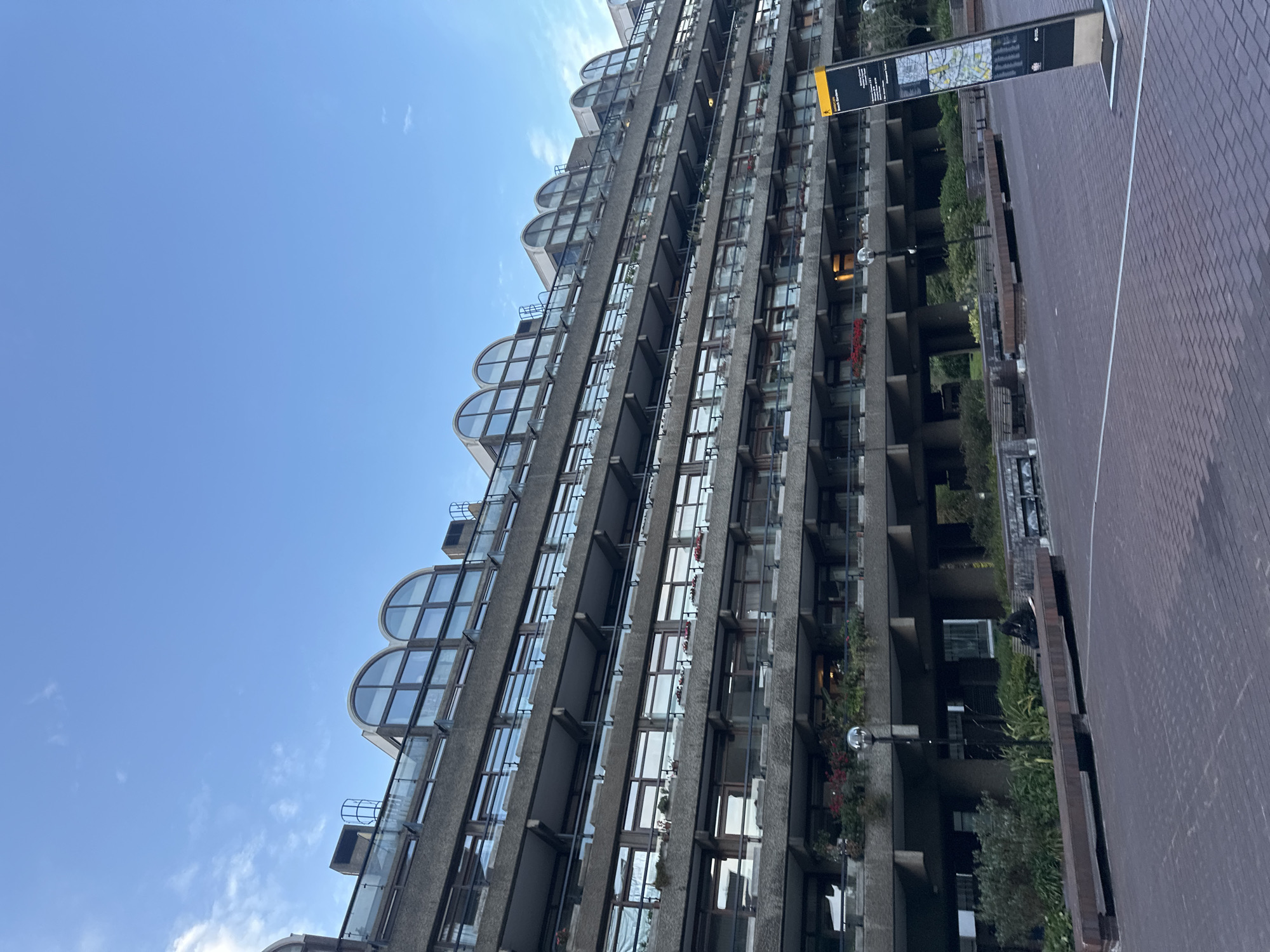 The photo shows a portion of the Barbican Estate in London, United Kingdom. The architecture features concrete structures, with multiple rows of apartments containing curved, semi-circular windows. Each apartment has a balcony with some greenery on the balconies. The sky is clear blue. A street runs alongside the building, paved with red brick. There is signage erected along the street.