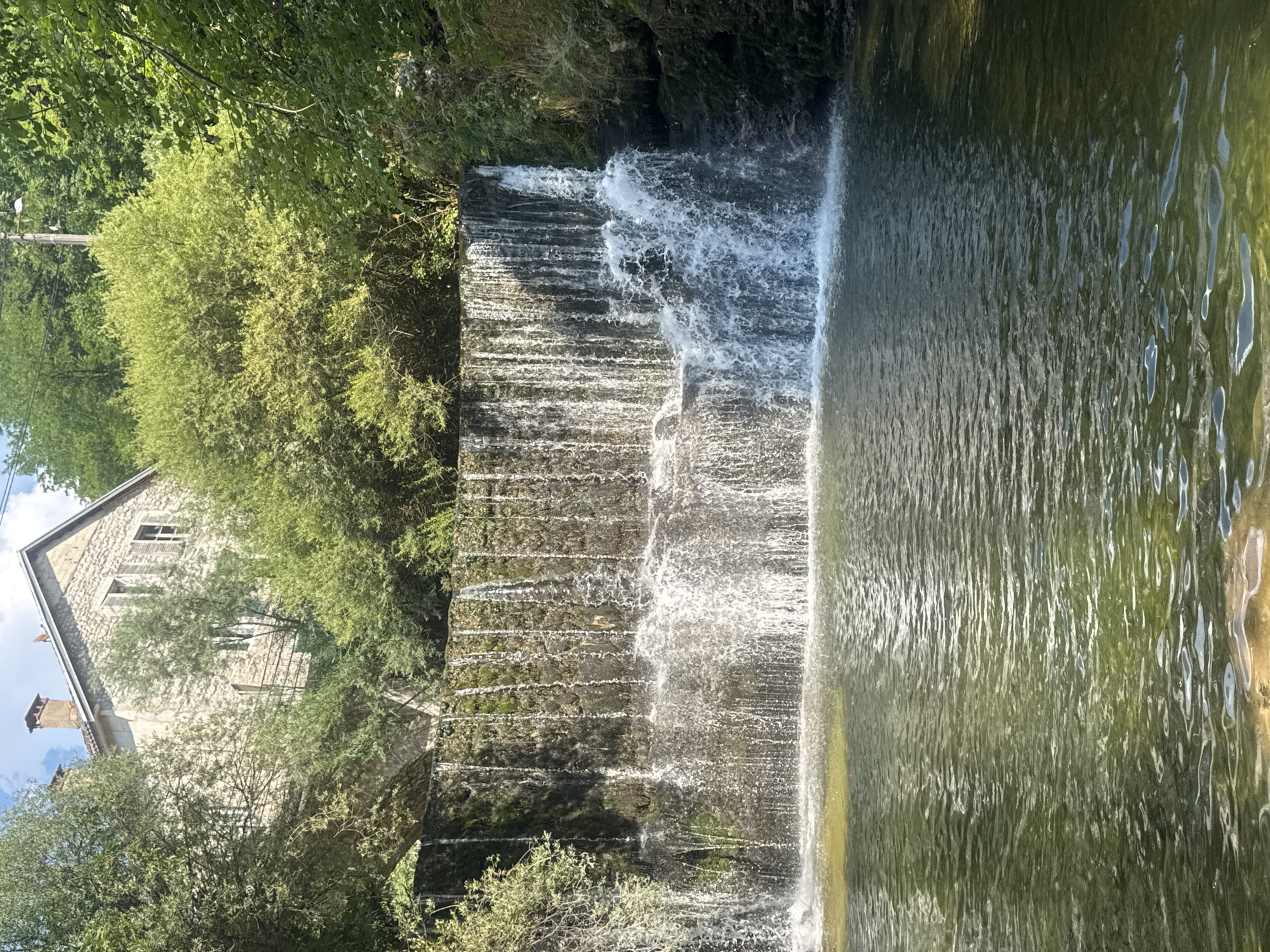 This media file displays a picturesque scene of a cascading waterfall in Saint-Claude, France. The waterfall, with its distinct tiered structure, flows over moss-covered stones, creating a textured and dynamic descent into a calm body of water. The water itself is a rich green hue, reflecting the lush vegetation surrounding it.
To the left of the waterfall, a quaint stone building is visible, nestled amongst abundant green trees and foliage. The building appears to be an older structure, possibly a house or a historical edifice, with windows and a chimney adding to its rustic charm. The presence of trees and greenery suggests a natural, possibly rural or semi-rural, setting. The sunlight filtering through the leaves indicates it is daytime, and the clear sky hints at pleasant weather conditions.
The overall impression is one of natural beauty and tranquility. The movement of the water and the static presence of the building and trees create a harmonious blend of nature and man-made elements. The image evokes a sense of peace and serenity, likely captured on a bright, clear day. There is no visible text in the media file.